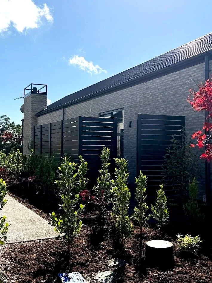 A modern house with gray brick walls and a black metal roof, featuring a chimney and a small deck with a privacy fence. There are landscaped plants and young bushes along a concrete walkway, with a clear blue sky overhead.