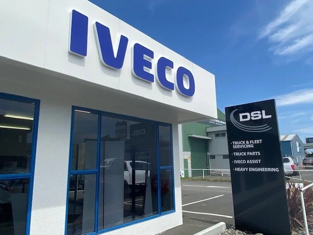 Iveco dealership with a large sign reading 'IVECO' and a smaller sign listing services like truck and fleet servicing, truck parts, Iveco Assist, and heavy engineering.