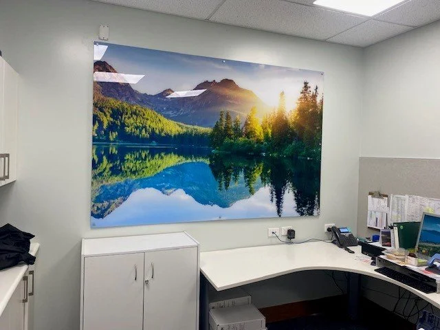 Office cubicle with a large landscape picture of mountains, a lake, and trees on the wall, along with office supplies and a computer.