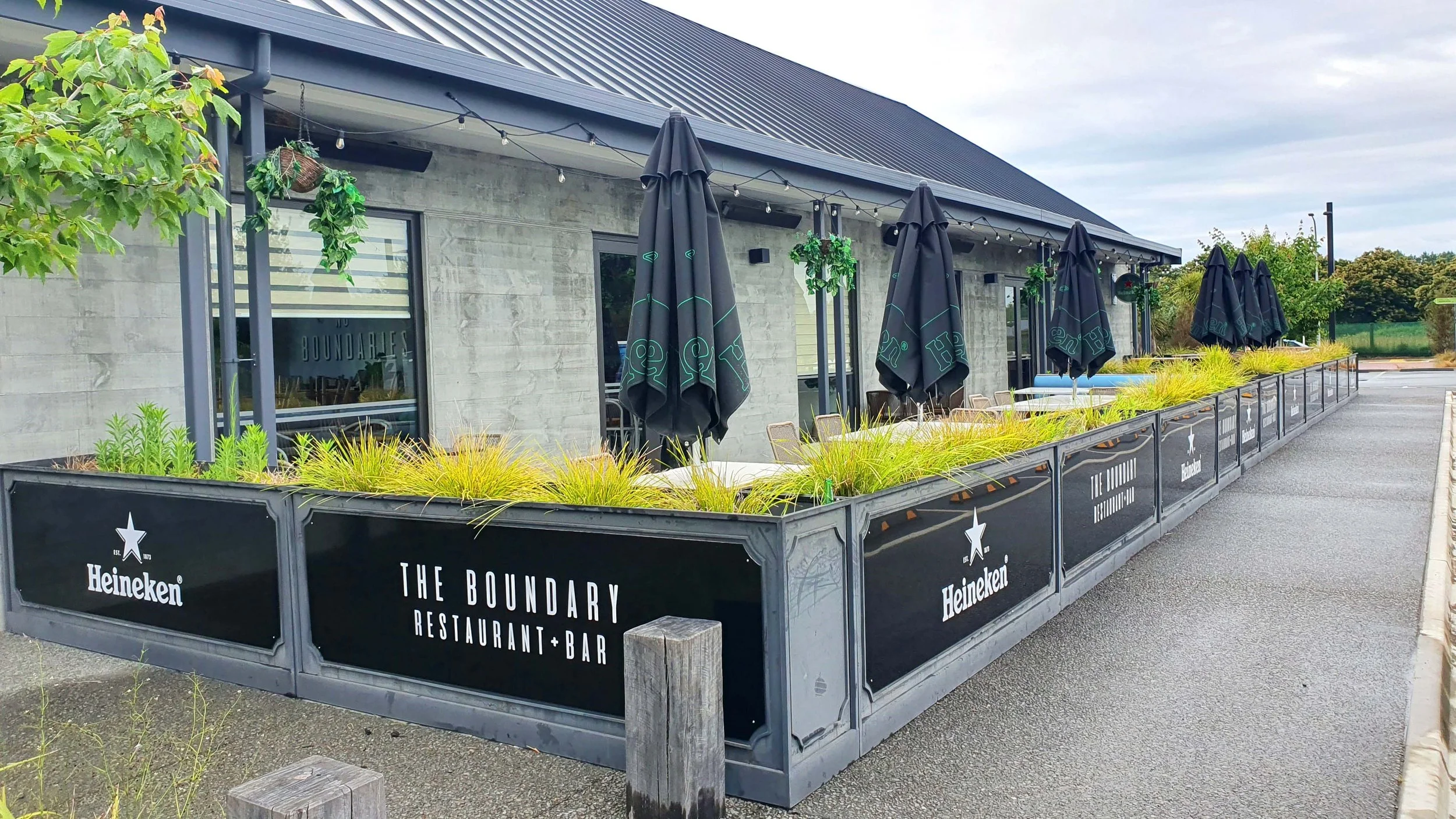 Outdoor patio of The Boundary restaurant and bar with black umbrellas, green plants, and a concrete building with large windows.