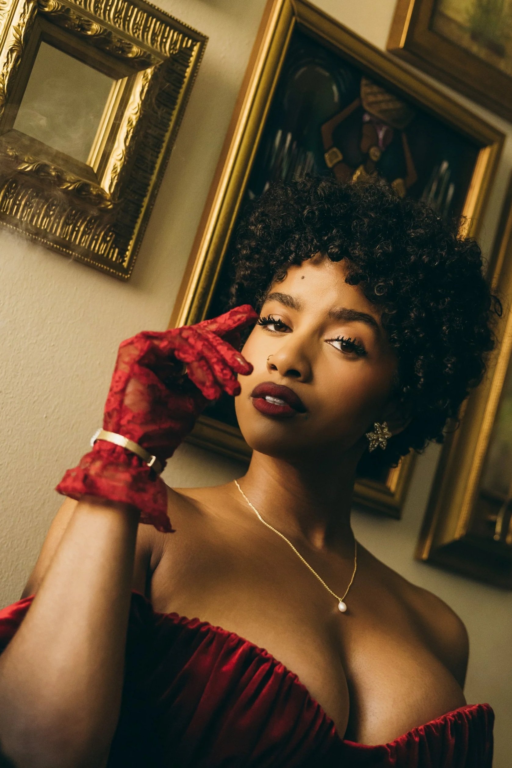 Ikea Damone , Singer on set  for her latest song called "Release"  rocking her curly hair, wearing red gloves, a strapless red dress, and jewelry, poses indoors against a wall adorned with framed pictures.