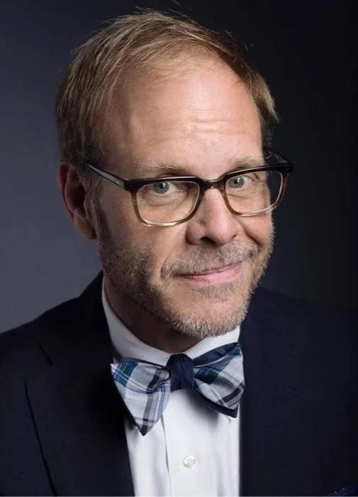 A collage of two photos of a middle-aged man with blonde hair and glasses, wearing a tuxedo and bow tie. In the left photo, he is holding a guitar covered with drawings and notes, with a white background. In the right photo, he is smiling against a d