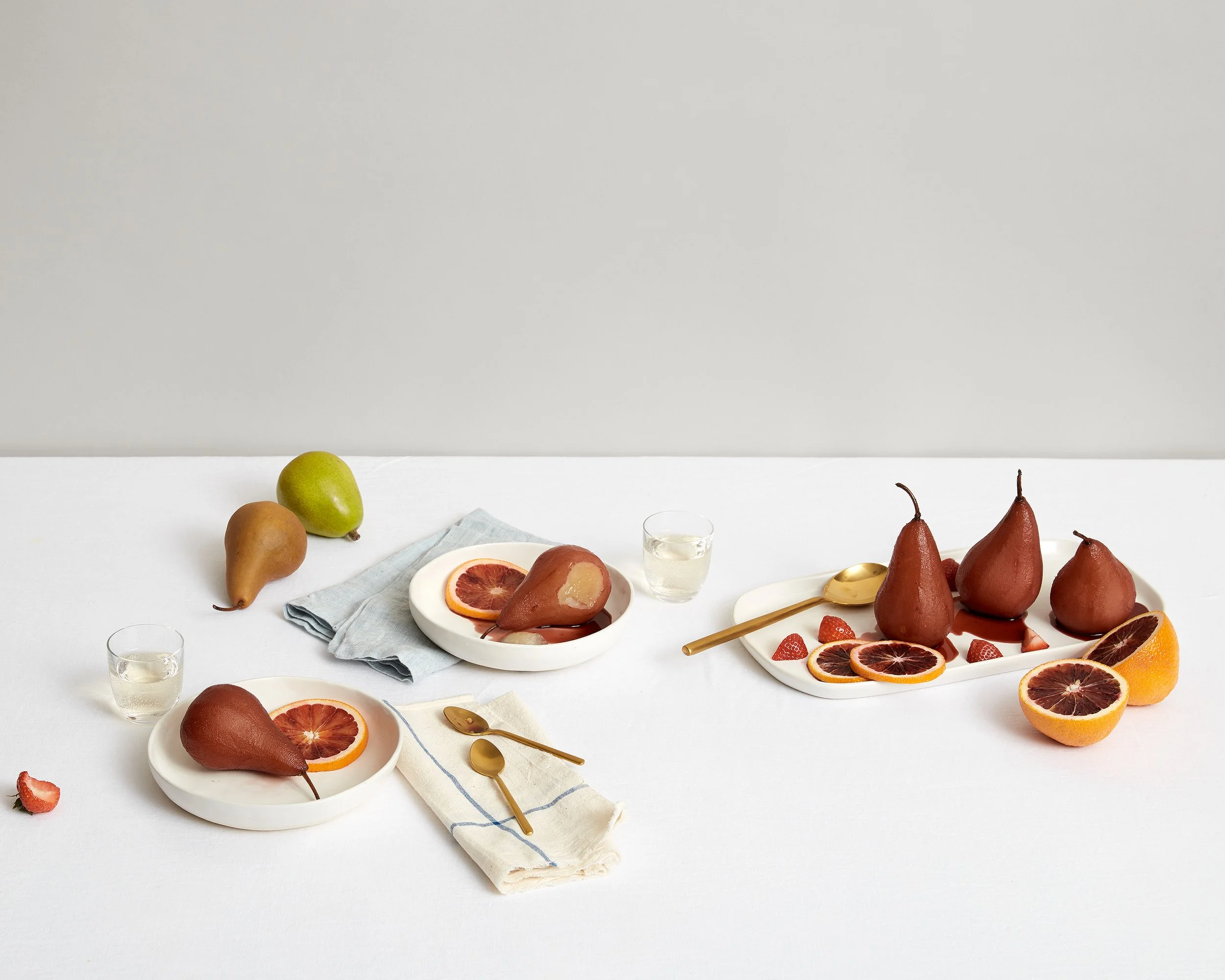 Decorative fruits, including pears, blood oranges, and halved oranges, arranged on white plates, with some fruits placed directly on the white surface and glasses of water in a minimalist setting.
