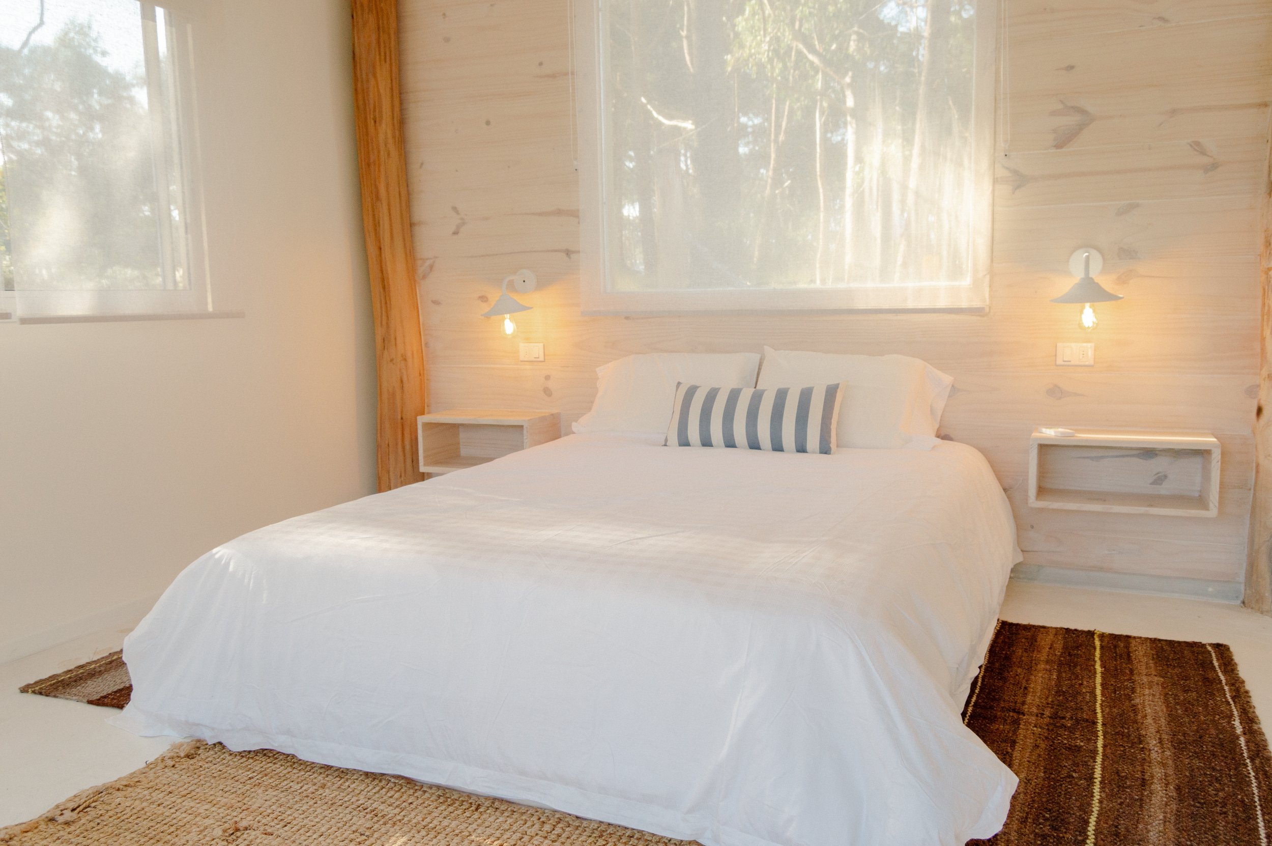 Modern bedroom with a white bed, striped pillow, wooden wall, bedside lamps, and window.