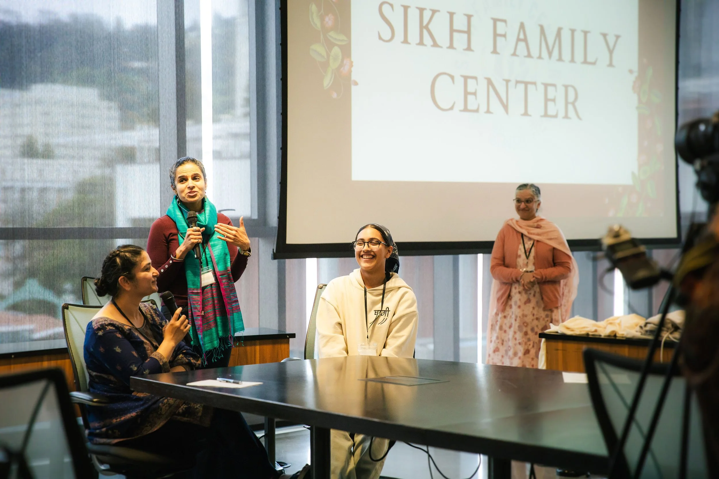 A group of five women in a conference room with large windows, one woman standing and speaking with a microphone, others seated and smiling, with a large screen displaying 'SIKH FAMILY CENTER' in the background.