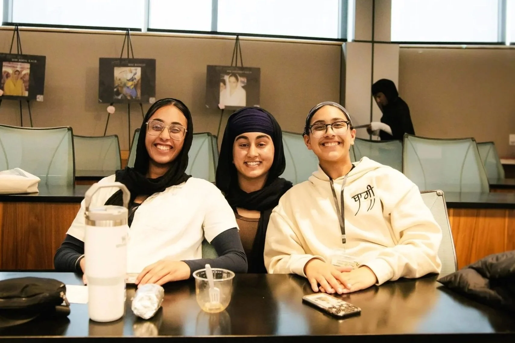 Three smiling Kaurs sitting at a table in a conference room with artwork on the wall behind them.