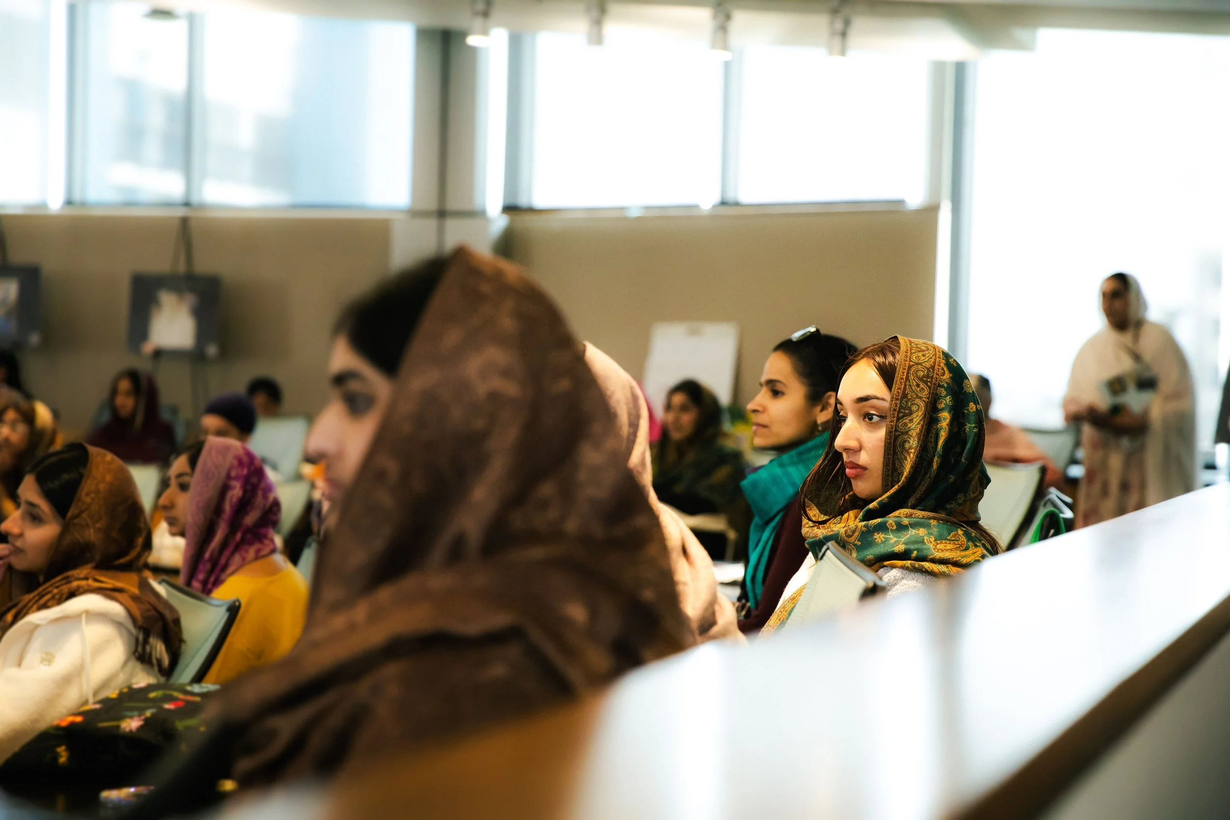 People attending a conference, sitting in chairs, some wearing headscarves, in a well-lit room with large windows.