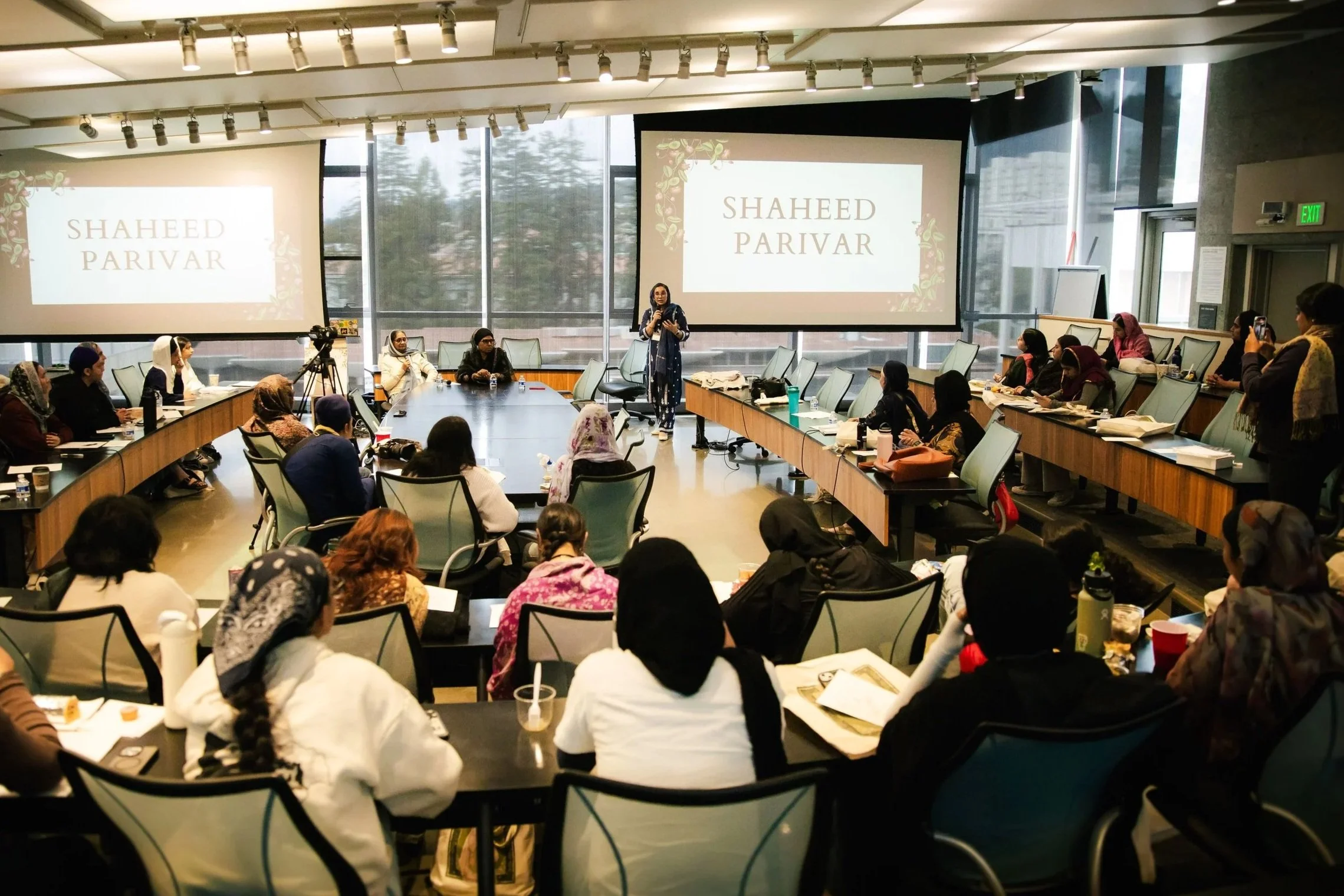 A conference room filled with people sitting around a U-shaped table, listening to a presenter standing in front of two large screens displaying the name 'Shaheed Parivar.' The room has large windows and a modern design.