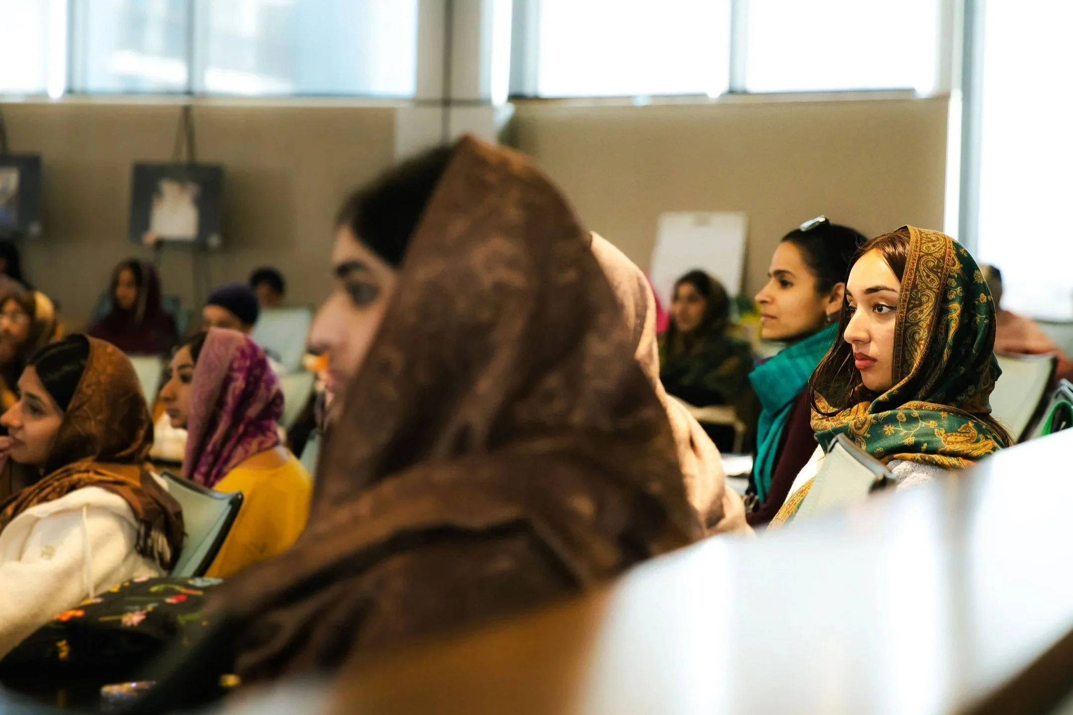 People attending a conference, sitting in chairs, some wearing headscarves, in a well-lit room with large windows.