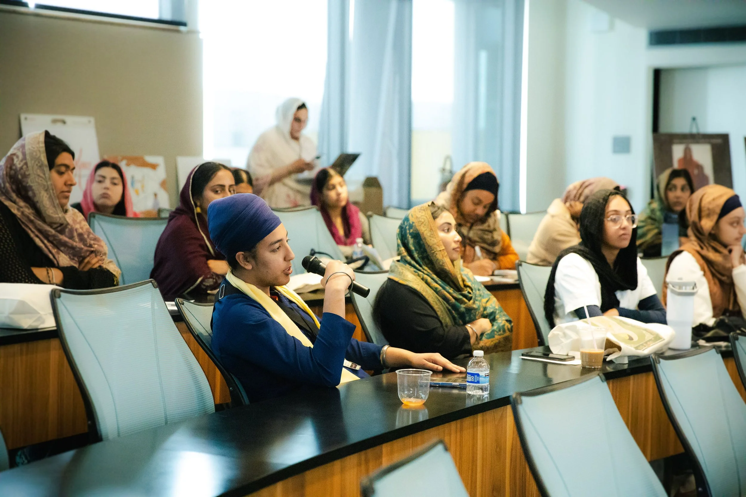 A diverse group of Sikh women, attending a conference or workshop in a room with large windows, some taking notes and one woman speaking into a microphone.