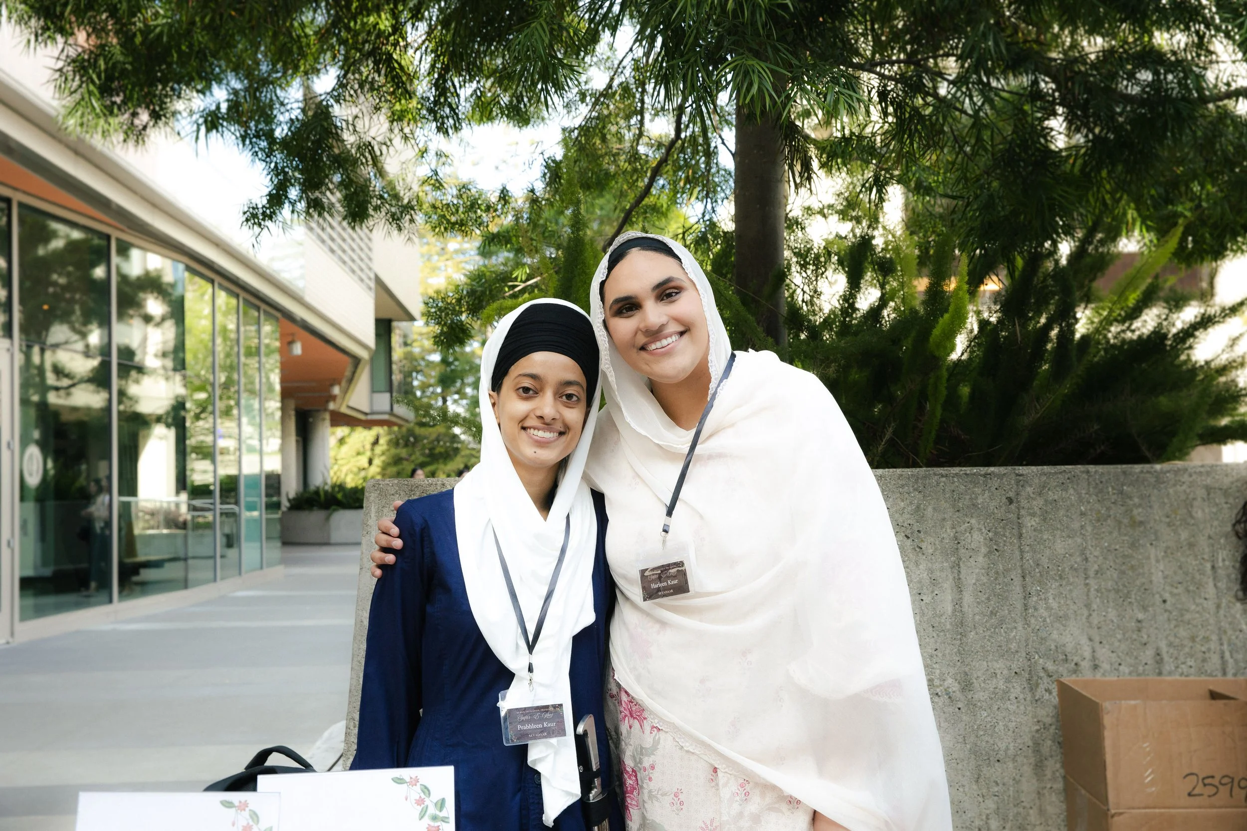 Two women wearing headscarves, smiling, standing outdoors near a concrete wall with greenery behind them. One woman is wearing a navy blue outfit, and the other is in a white outfit. They have name tags hanging around their necks.