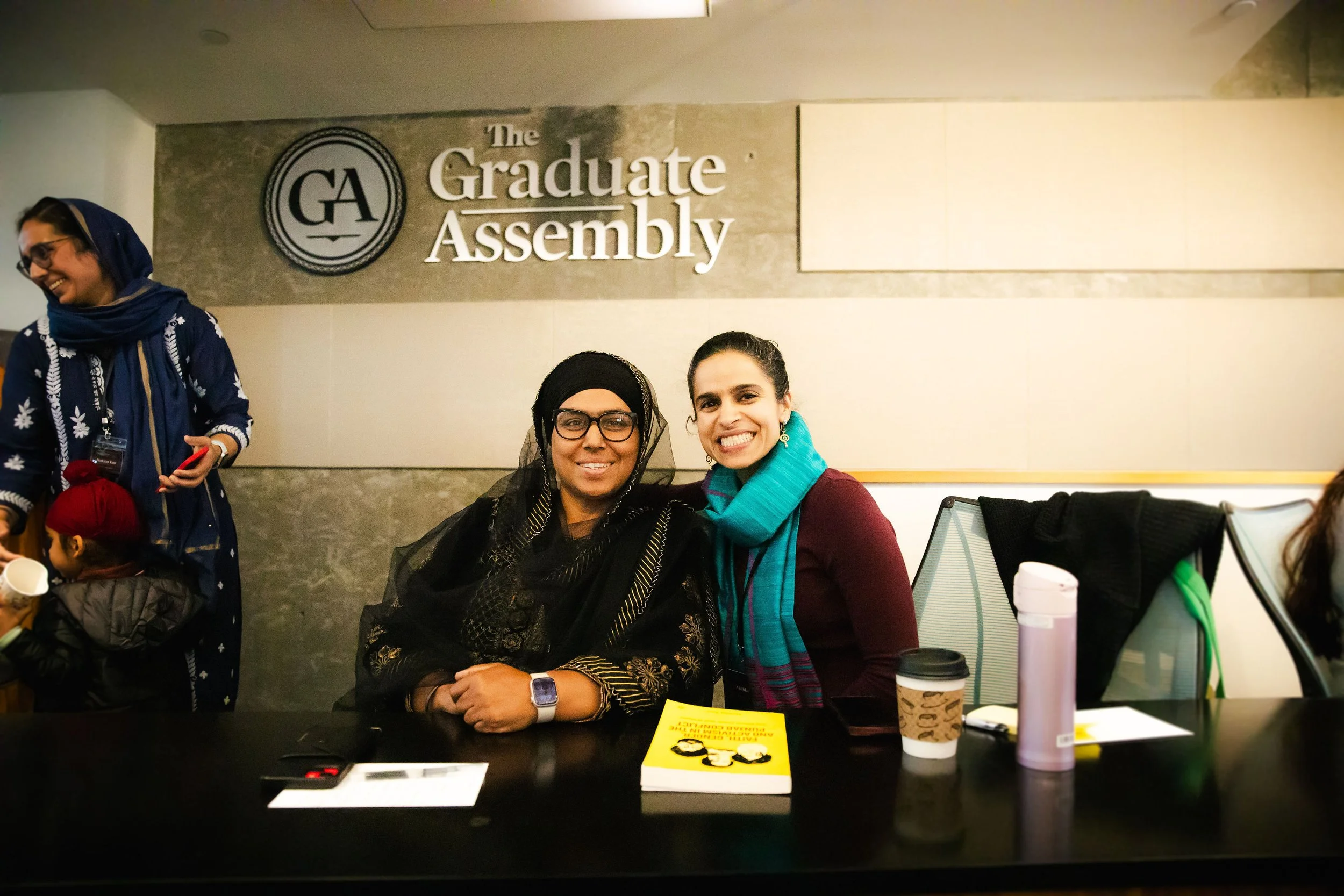 Two women sitting at a table smiling, with a sign reading 'The Graduate Assembly' in the background.