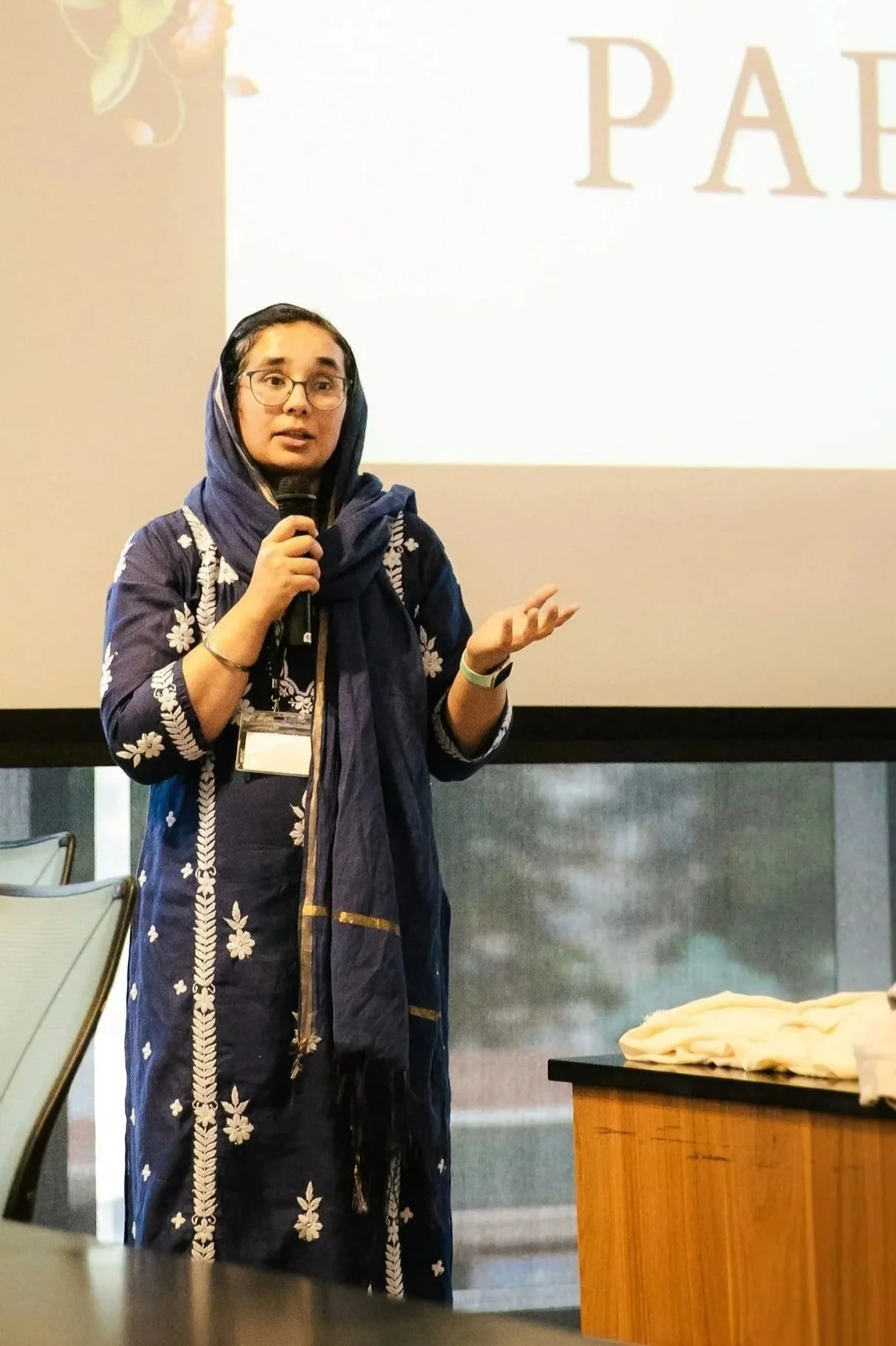 Bibi Navkiran Kaur, Jaswant Singh Khalra's daughter, in traditional Punjabi attire giving a presentation with a microphone in front of a projection screen.