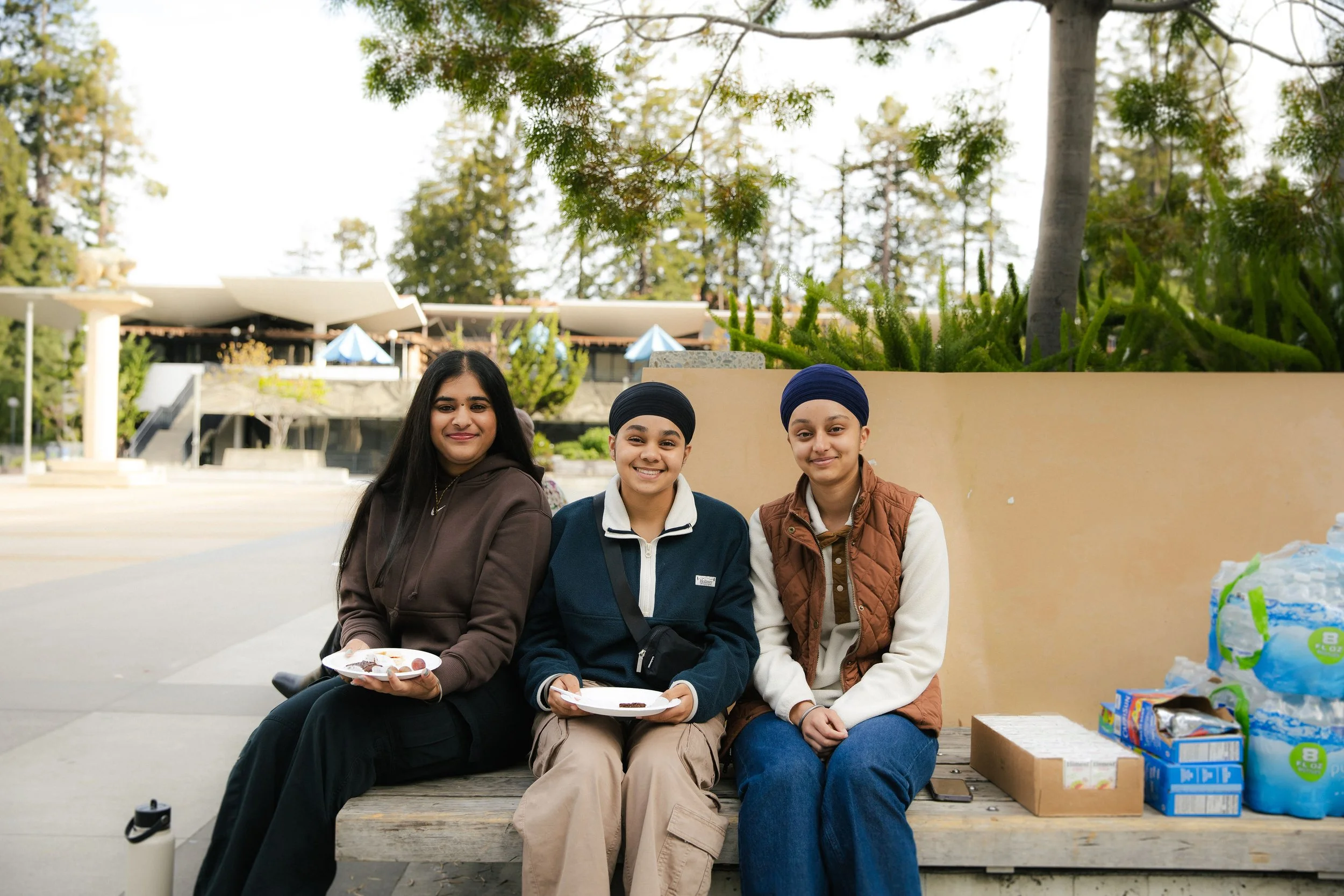 Three young women sitting on a bench outdoors, smiling and holding plates of food, with supplies on the right side and greenery behind them.