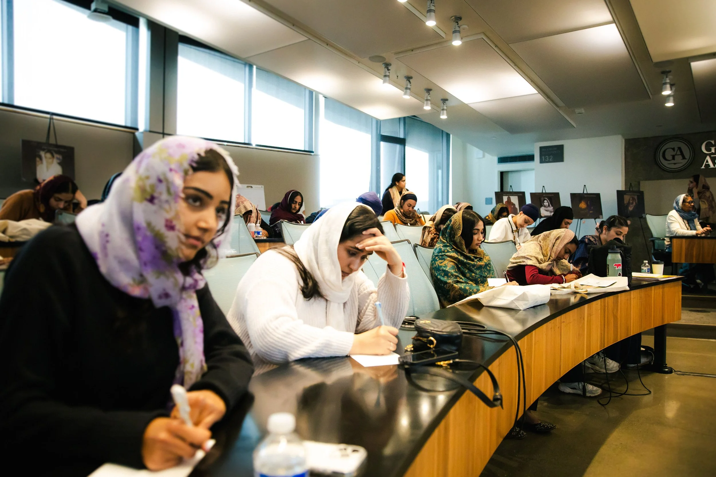 A diverse group of women attending a class or seminar in a large, modern conference room, seated at curved tables and taking notes.