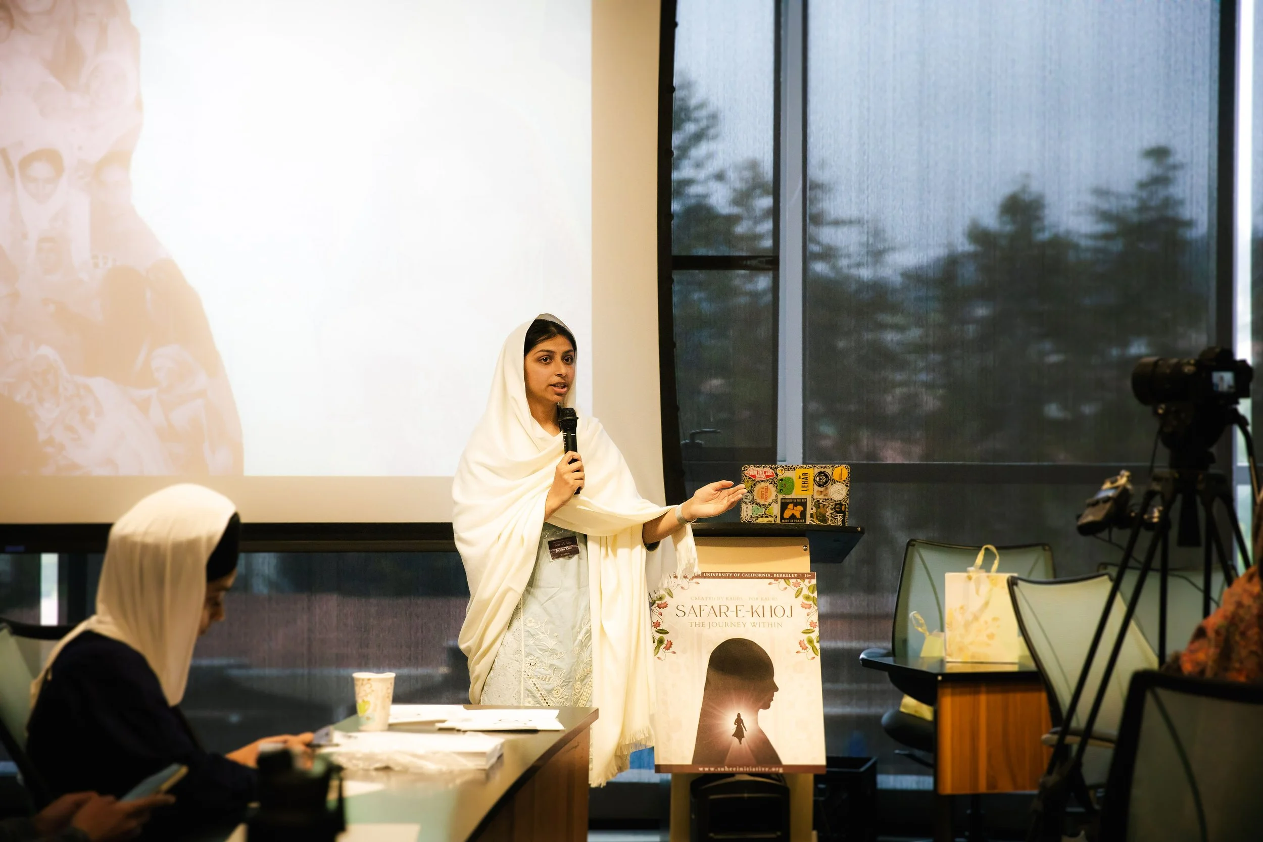 A woman dressed in white is giving a presentation in a conference room, standing near a screen and a poster that reads "SAFAR-E-KHOJ: THE JOURNEY WITHIN". She is holding a microphone and gesturing with her other hand. There are people seated at table