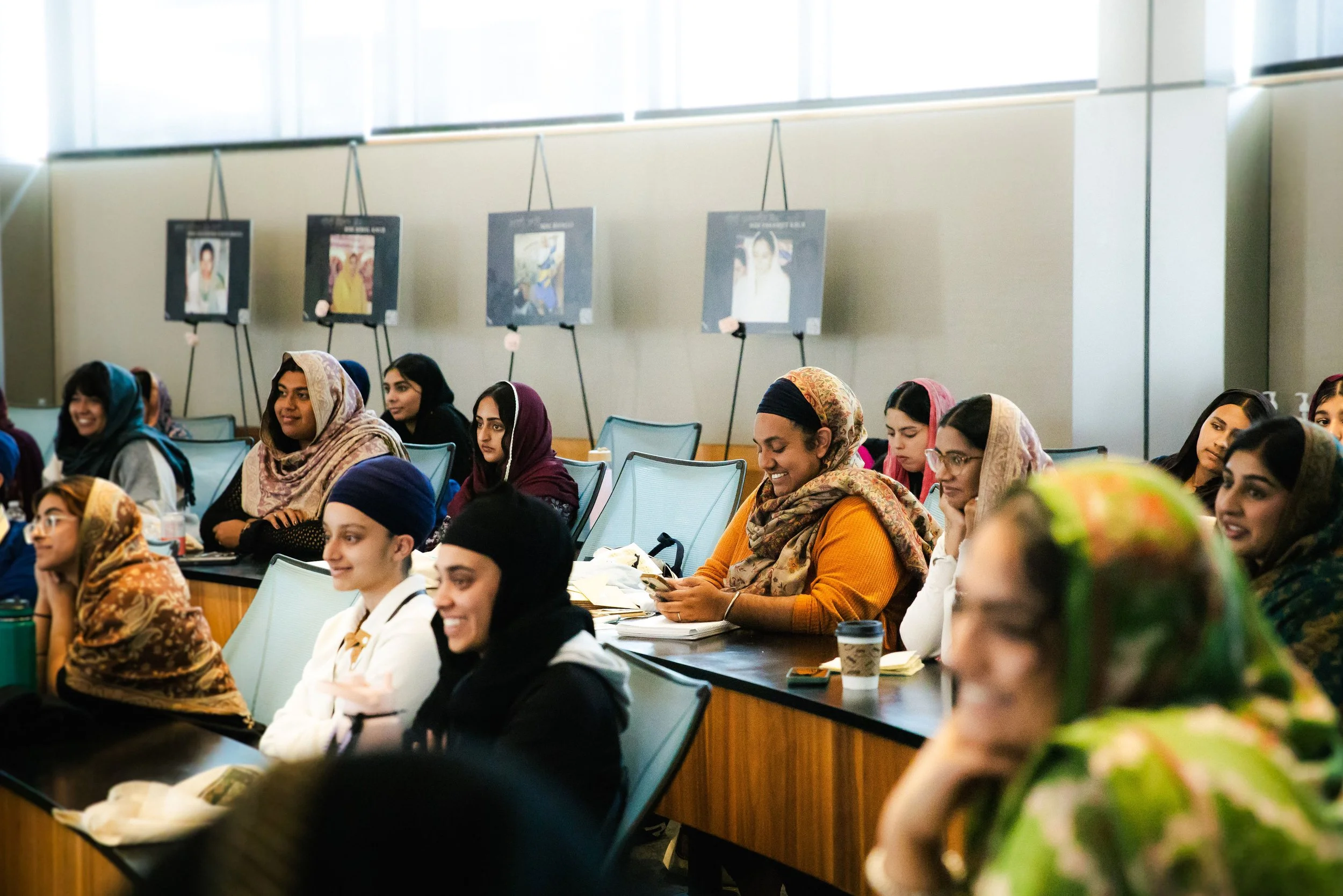 Group of women and girls sitting and smiling at an indoor conference or seminar, some wearing colorful headscarves, with portraits on display in the background.