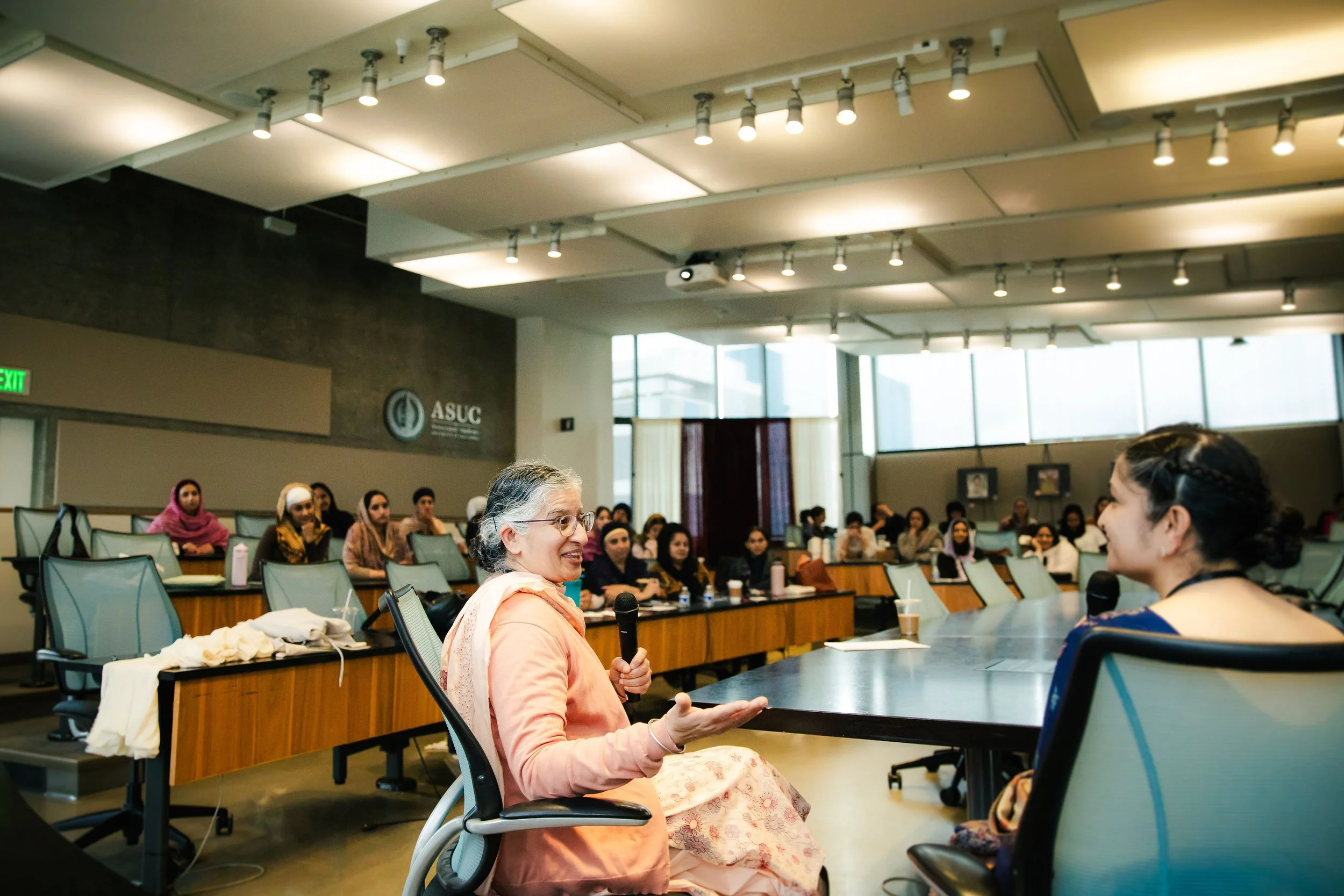 A diverse group of Kaurs Sikh women attending a seminar in a large conference room with natural light, with one woman from Sikh Family Center speaking into a microphone.
