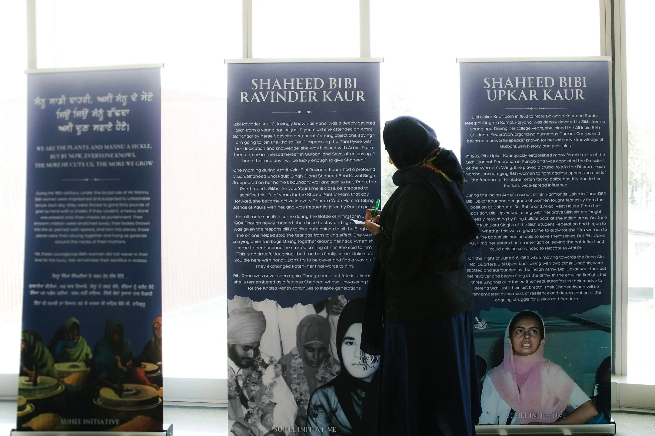 A woman in dark clothing and a head covering is reading from a framed display of informational posters about Shaheed Bibi Ravinder Kaur and Shaheed Bibi Upkar Kaur, with additional handwritten and printed text in the background.