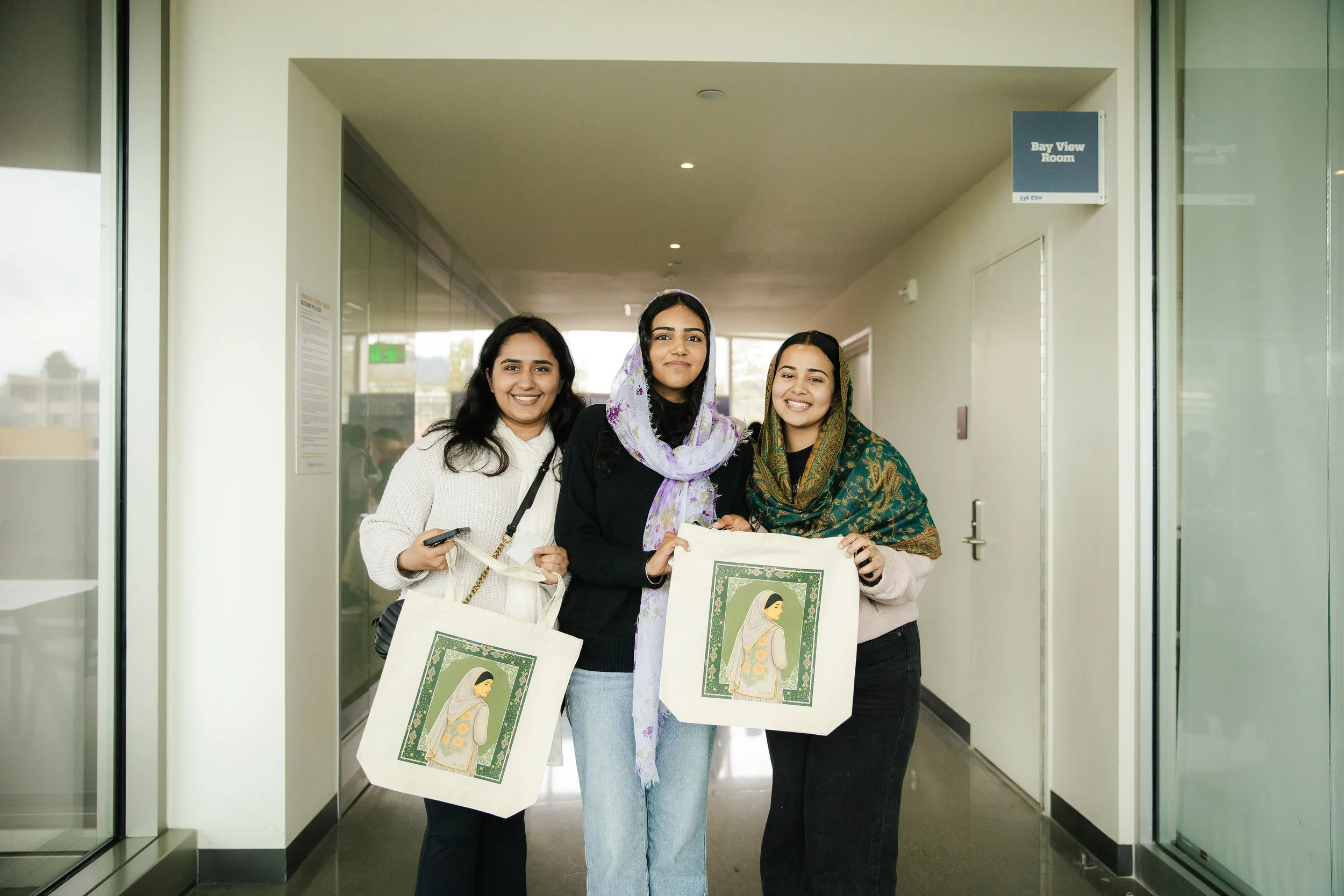 Three smiling women standing in an indoor hallway, holding tote bags with an artwork of a woman in traditional attire, with a background of a window showing a parking lot.