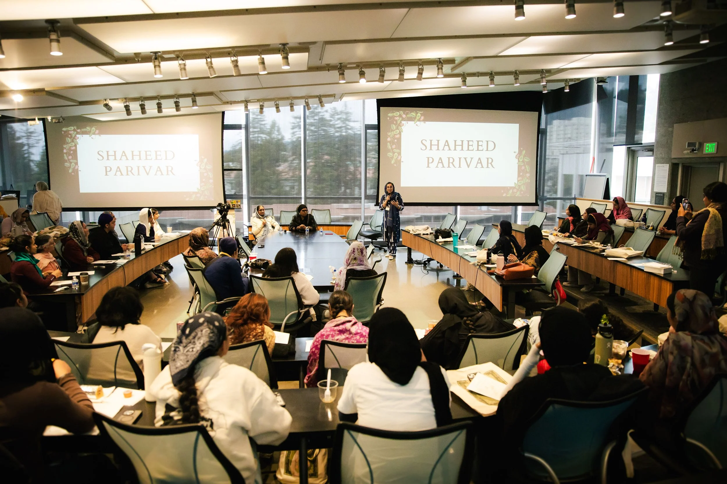 A conference room filled with people sitting around a U-shaped table, listening to a presenter standing in front of two large screens displaying the name 'Shaheed Parivar.' The room has large windows and a modern design.