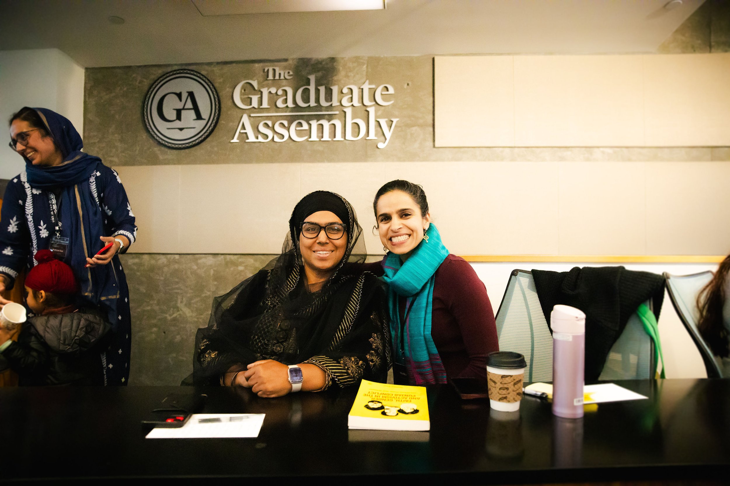 Two women sitting at a table smiling for the camera in front of a sign that reads "The Graduate Assembly" at Georgia Tech.