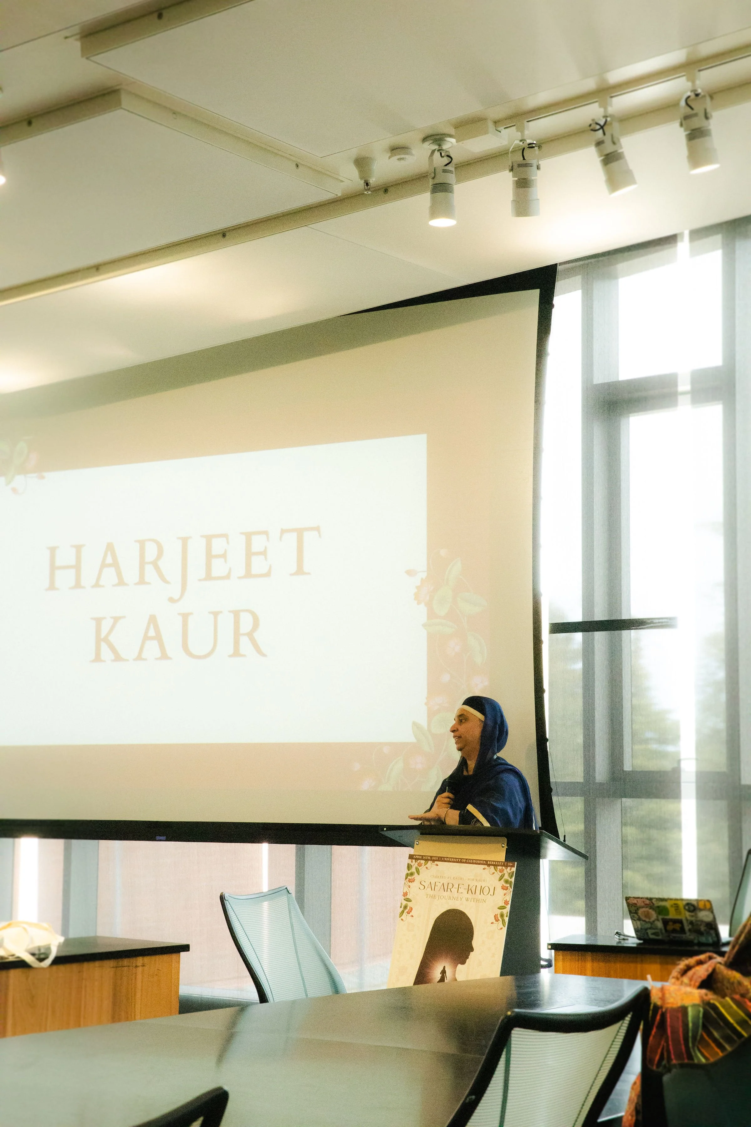 A woman dressed in traditional South Asian attire, standing at a podium, delivering a presentation. A large screen behind her displays the name 'Harjeet Kaur' with floral decoration. The room has large windows, and there are chairs and tables in the 