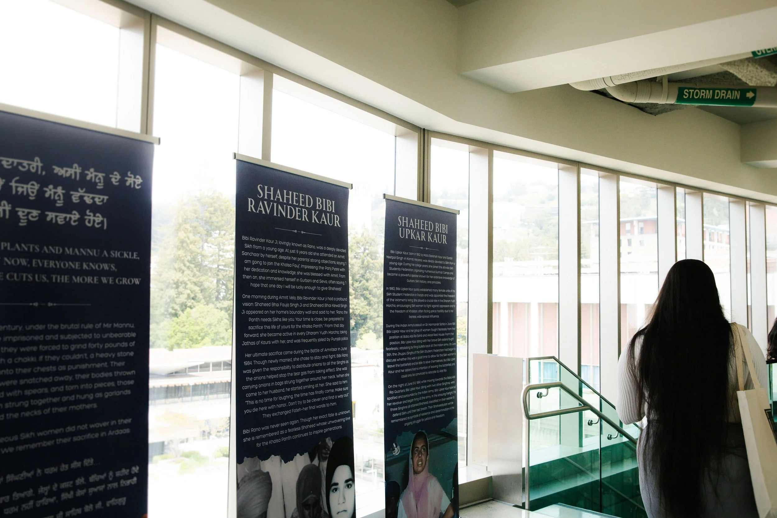 A woman viewing informational display banners inside a building near large windows with outdoor trees and buildings visible.