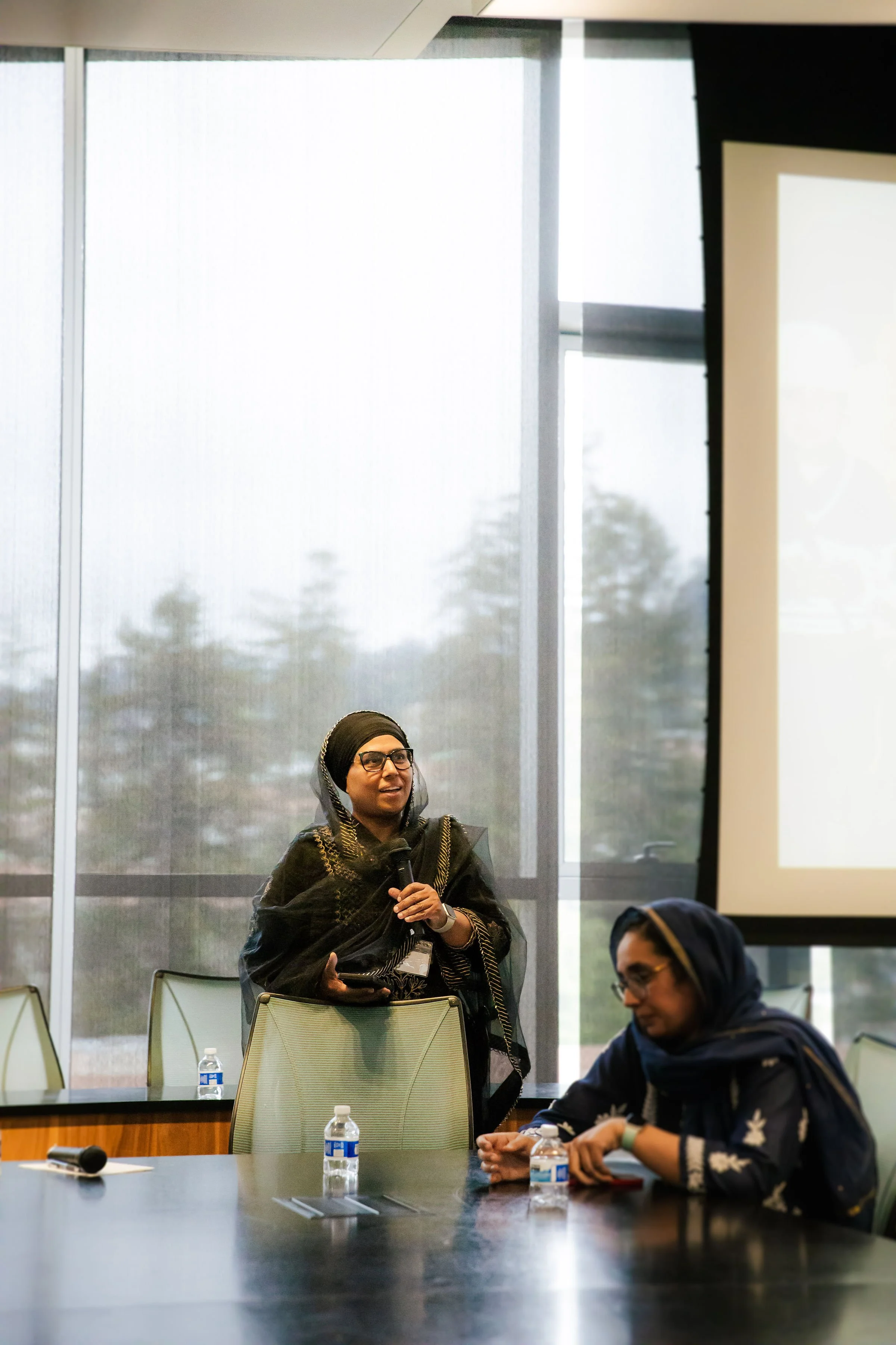 Two women in headscarves are in a conference room with large windows, one standing and speaking into a microphone and the other sitting with bottled water in front of her.