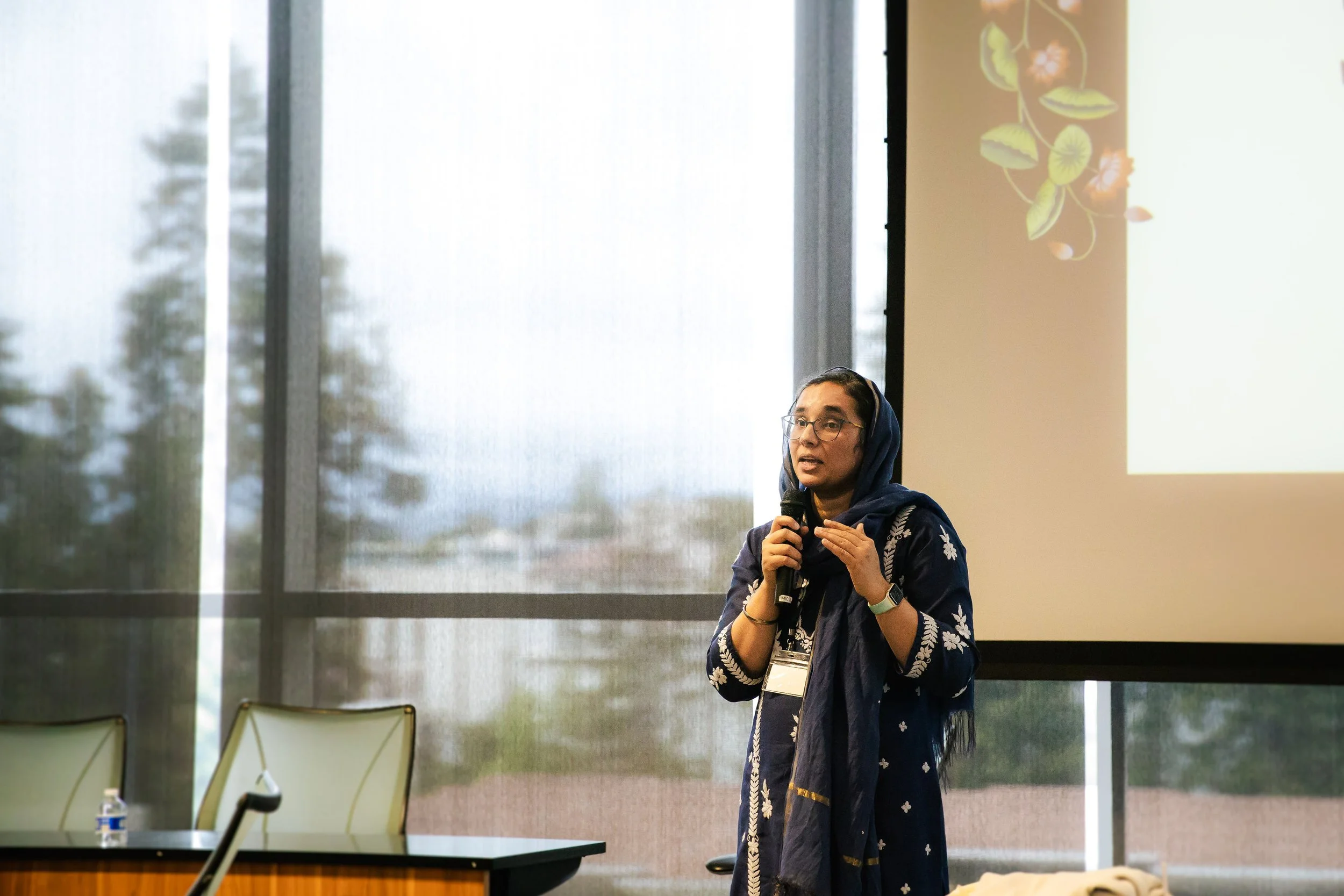 Woman in traditional South Asian attire speaking into a microphone during a presentation in a conference room.
