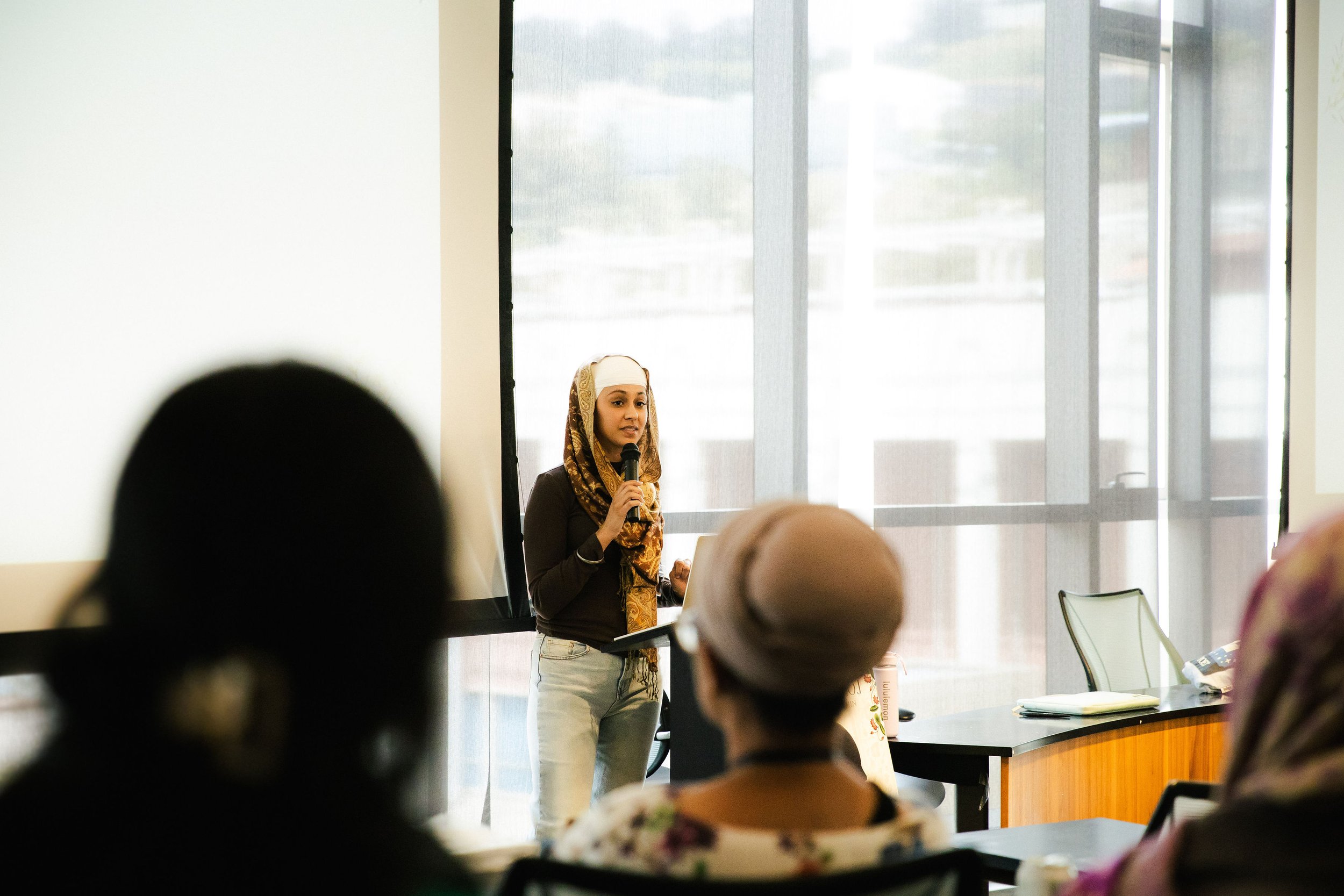 A woman wearing a headscarf and dark top giving a presentation in front of a room of people, holding a microphone.