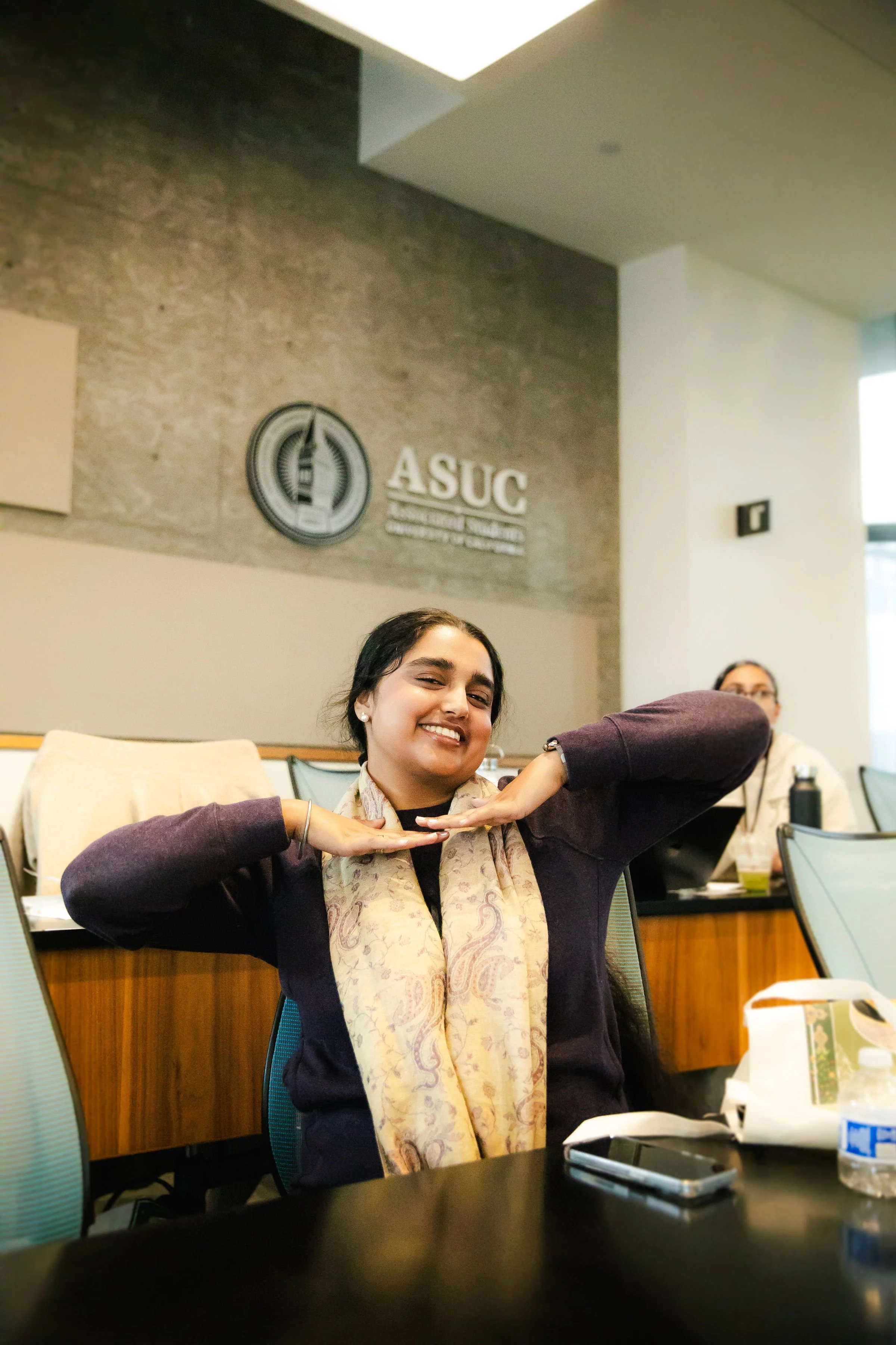 Smiling woman with dark hair in a conference room making a playful gesture with her hands under her chin, with a woman with glasses in the background at a table with papers and a water bottle, in front of a wall with ASUC sign at UC Berkeley.