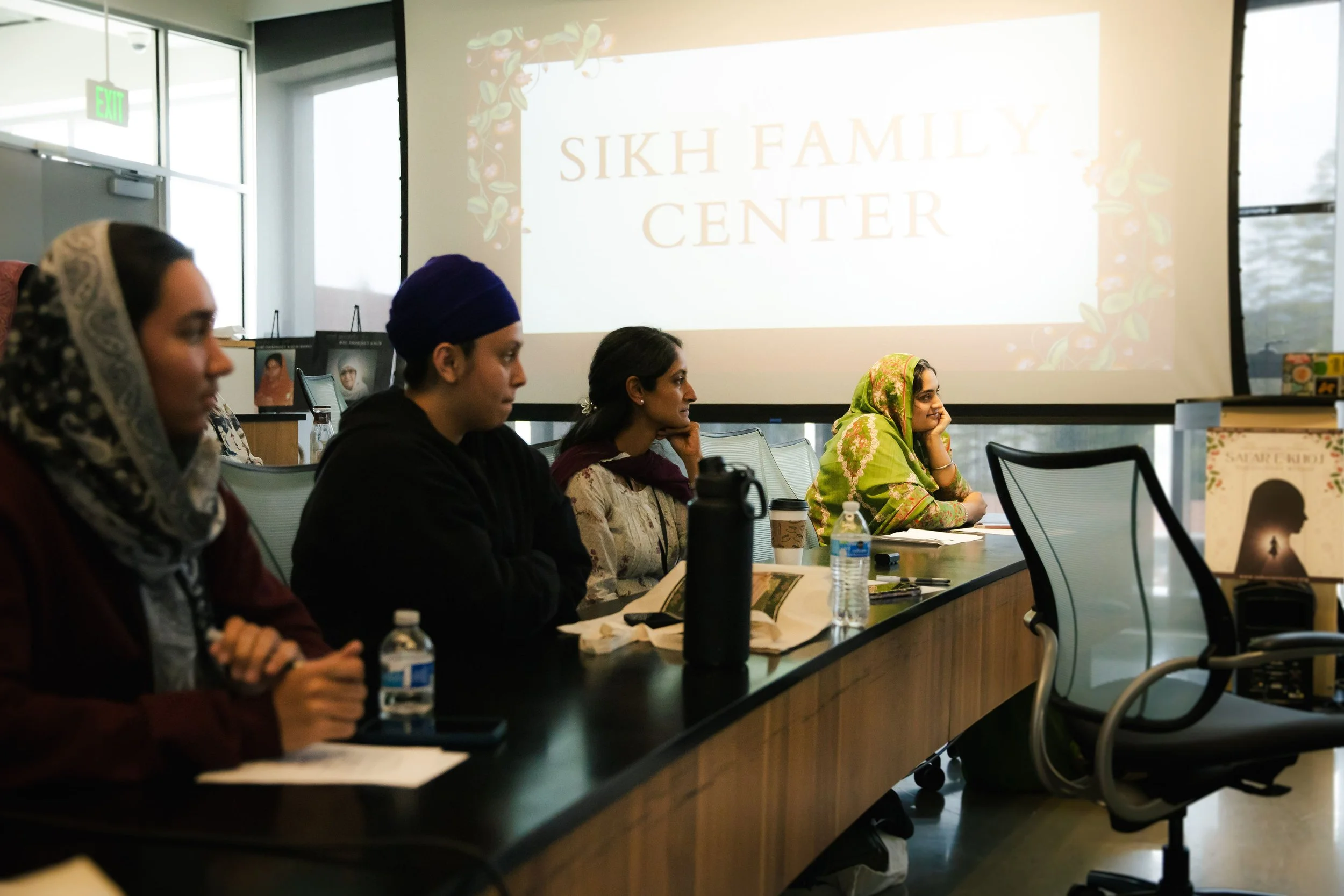 Women of Sikh heritage attending a meeting at the Sikh Family Center, sitting at a conference table with water bottles and notebooks, in a room with large windows and a projector screen displaying the center's name.