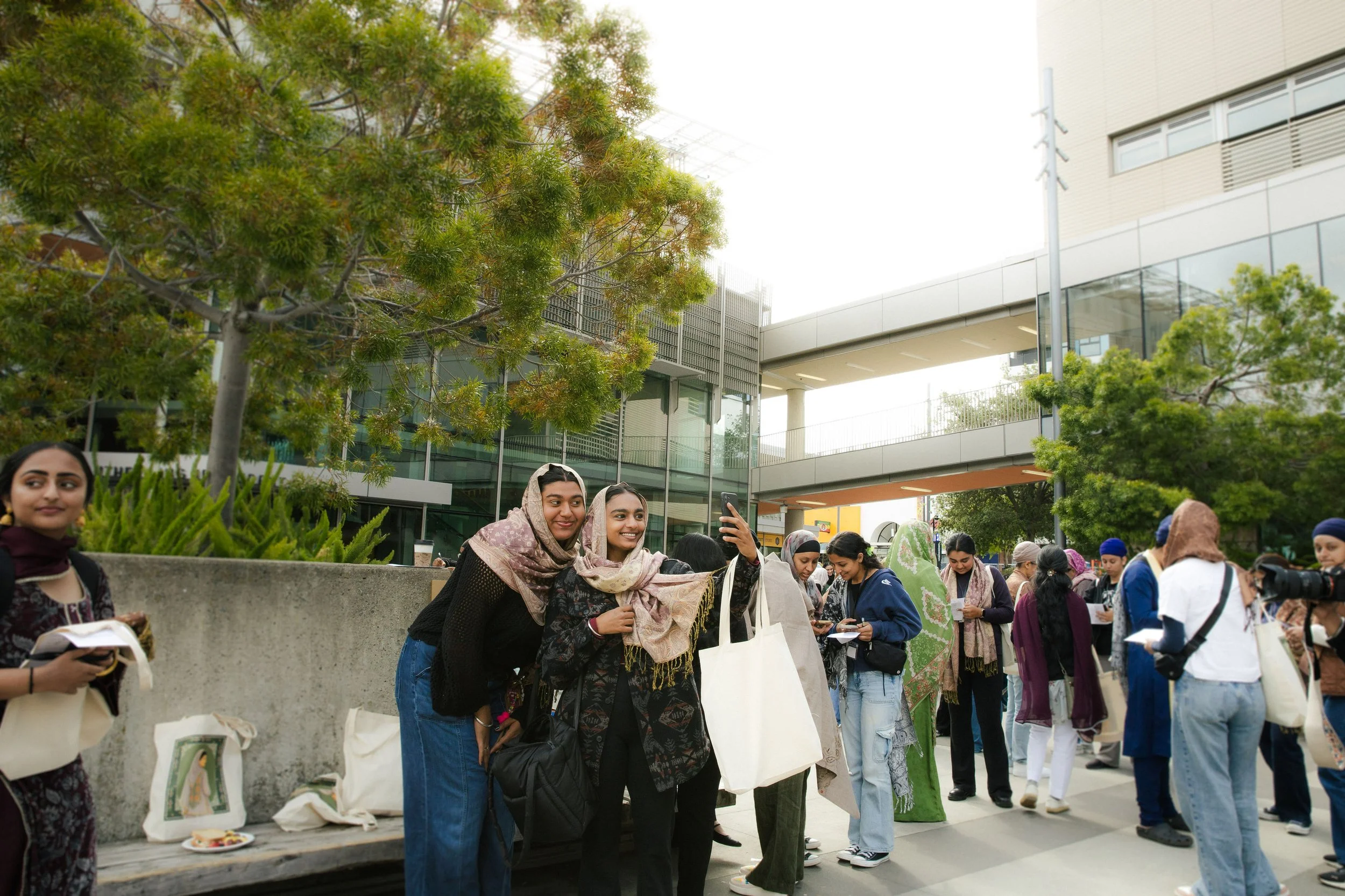 Group of people gathering outdoors near modern buildings, some taking photos, others holding papers or bags, with trees and a bridge in the background.