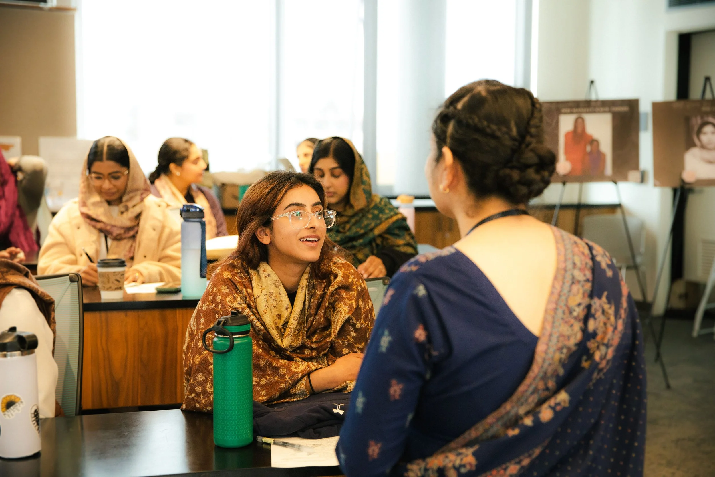 Women engaging in conversation in a classroom setting with posters on easels in the background.