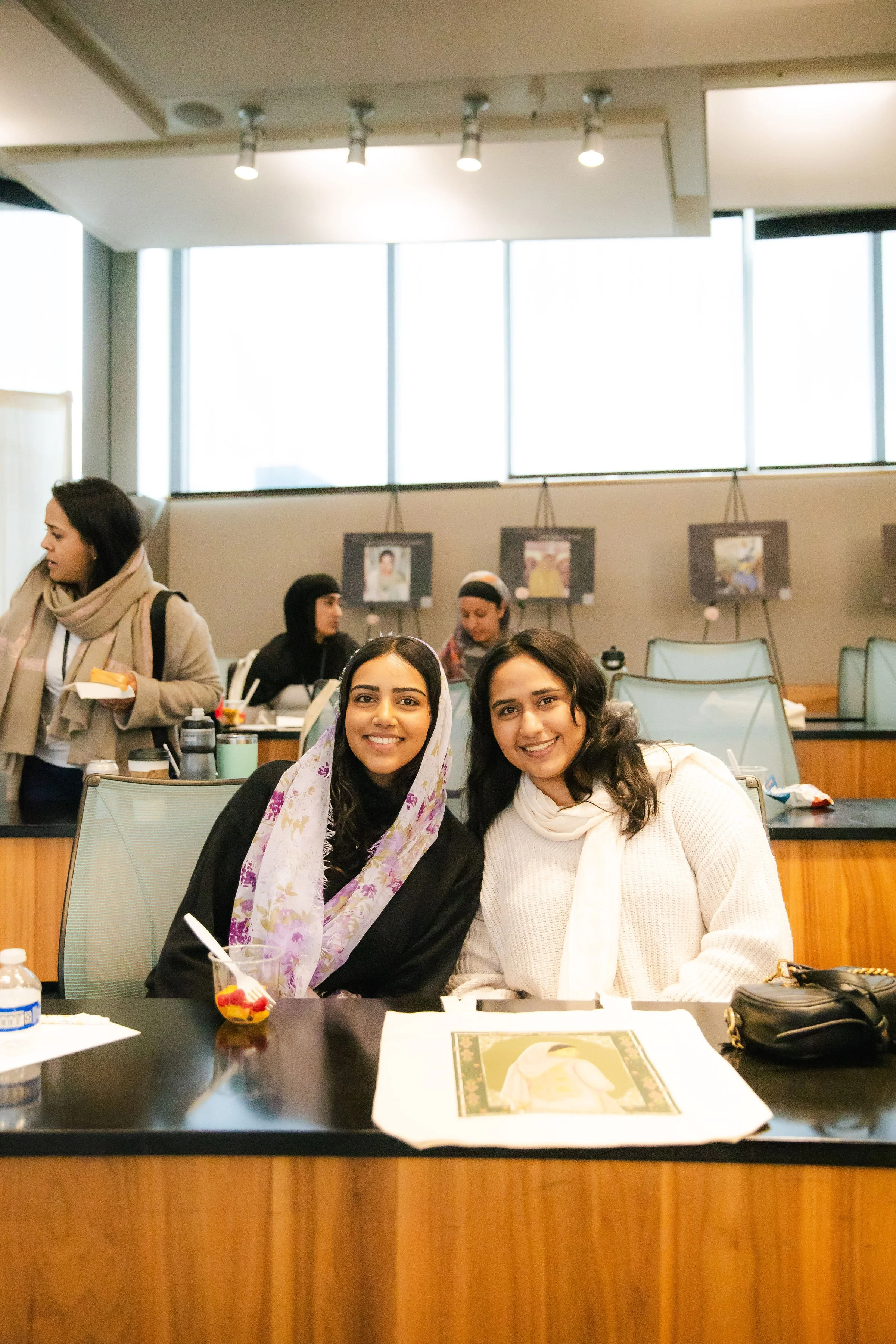Two women sitting at a table in a cafe or restaurant, smiling at the camera, with artwork on the wall behind them and other people in the background.