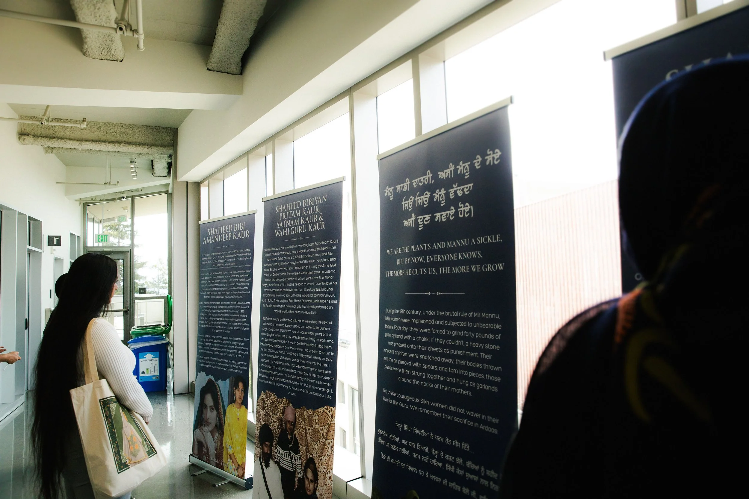 People reading informational display panels in an indoor gallery with large windows and overhead pipes.
