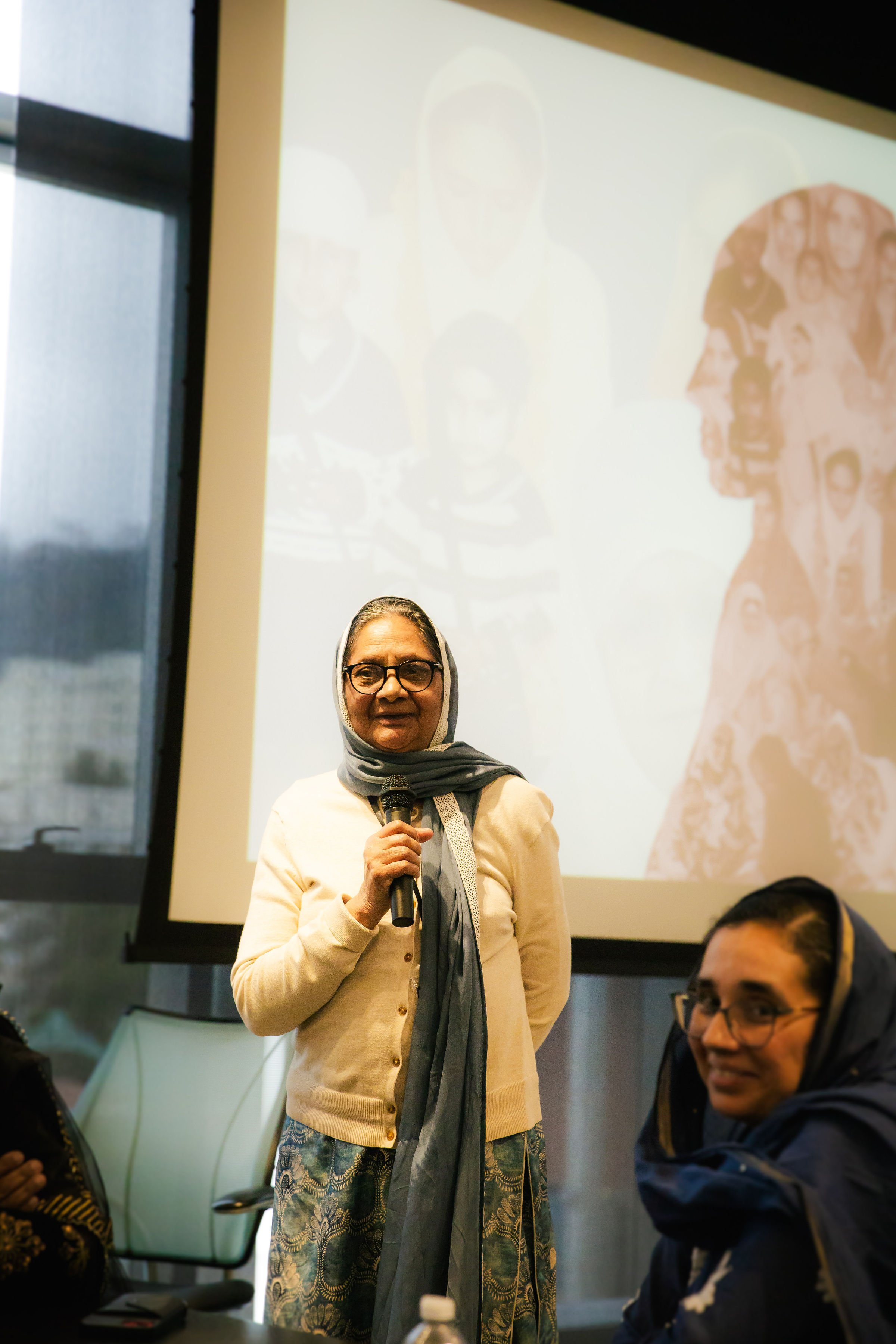  with glasses and a grey headscarf holding a microphone, standing in front of a large screen displaying a collage of photographs of children and adults. A man wearing glasses and a blue hoodie sits nearby, smiling. The setting appears to be a confere