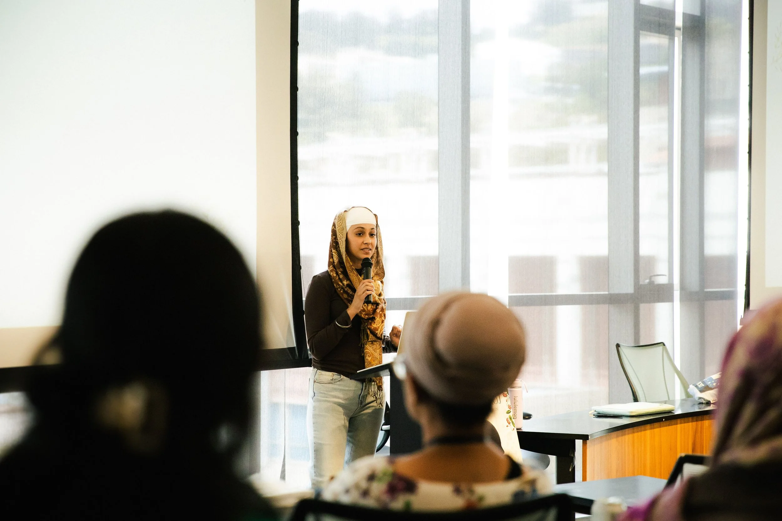 A woman in a hijab holds a microphone and speaks to an audience in a conference room with large windows.