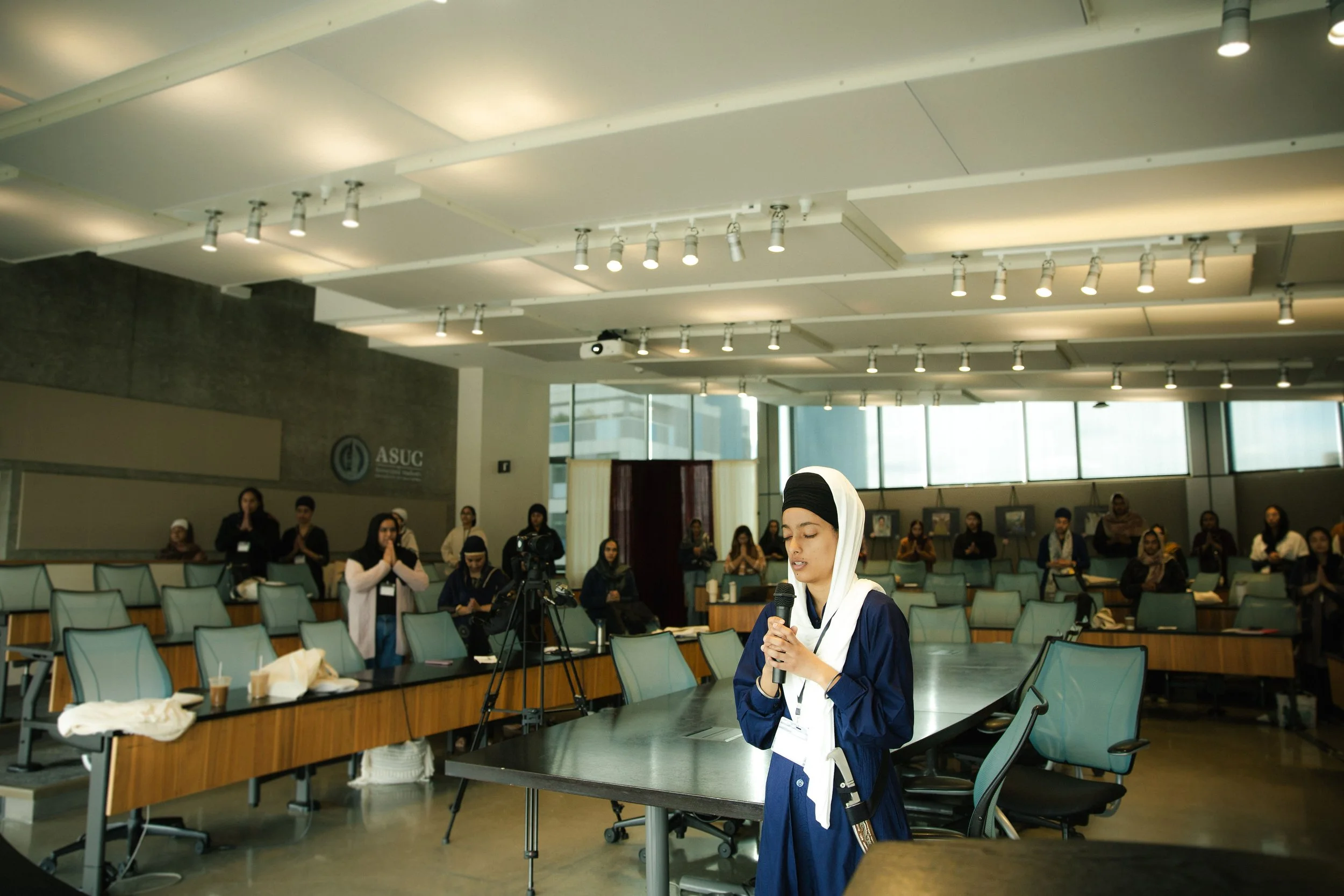 A young woman in a blue dress and white headscarf standing at a table with a microphone, speaking or praying, in a conference room with several people standing and seated behind her.