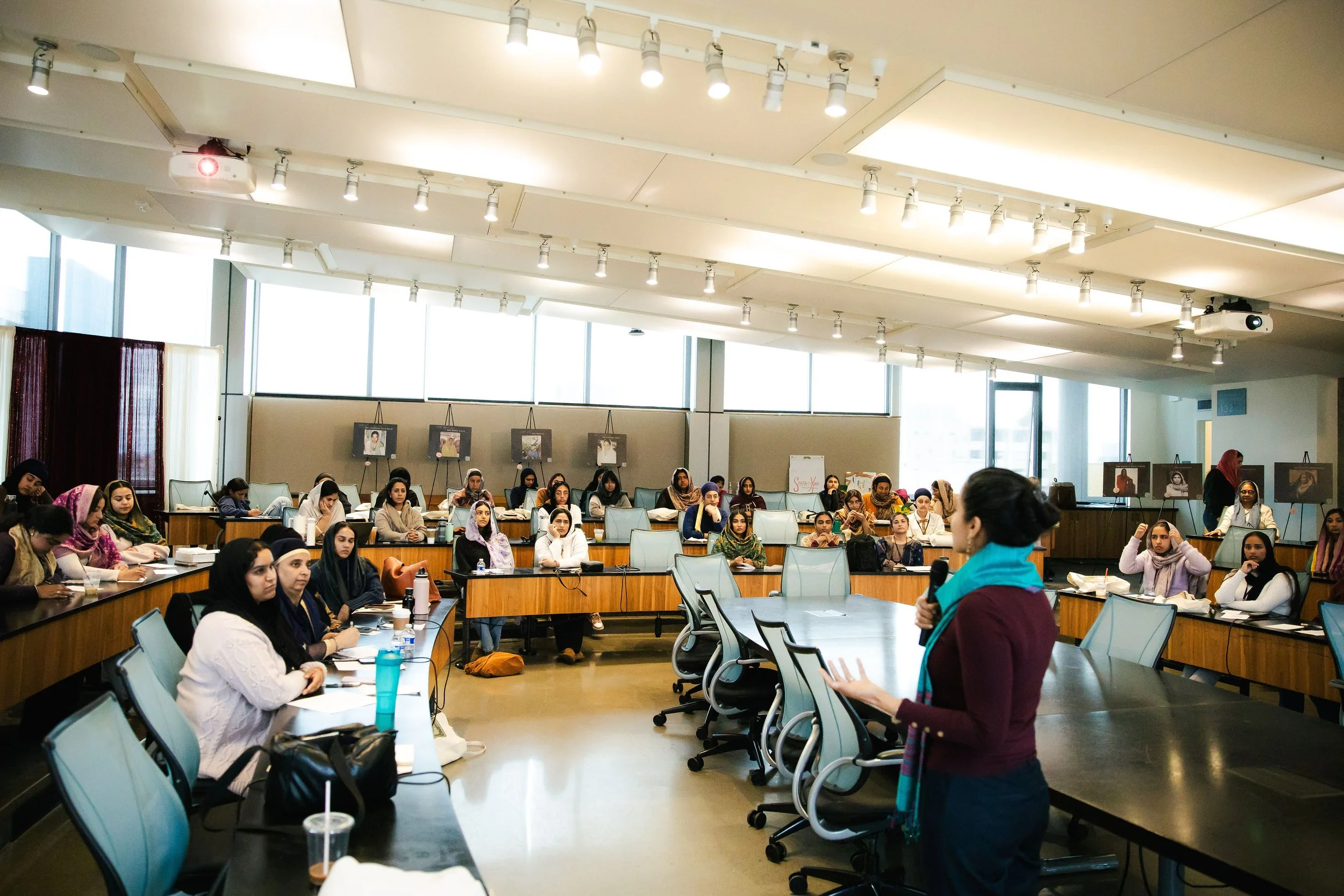 A woman giving a presentation to a large group of women seated around a U-shaped conference table in a well-lit room with large windows and framed photographs on the wall.