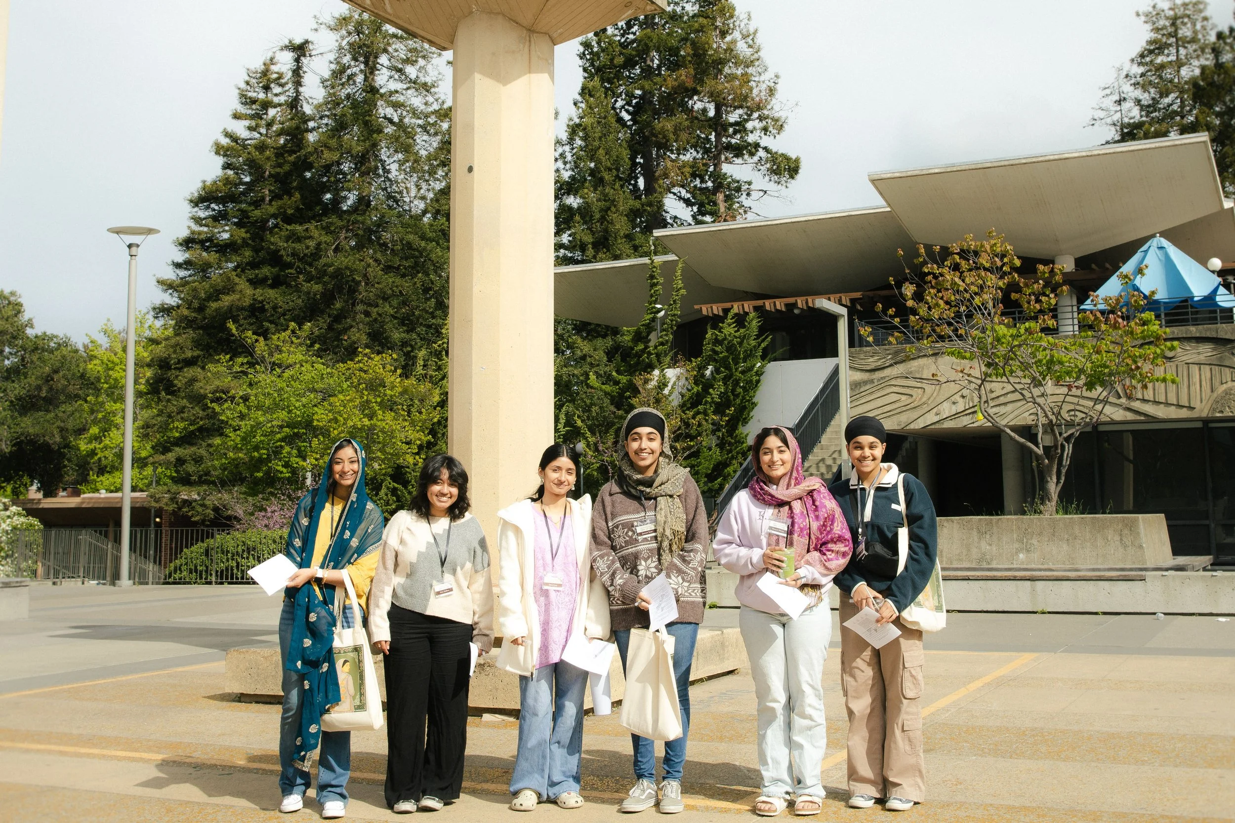 Group of six diverse young women standing outdoors in front of a modern building, smiling and holding papers and tote bags, with trees and greenery in the background.