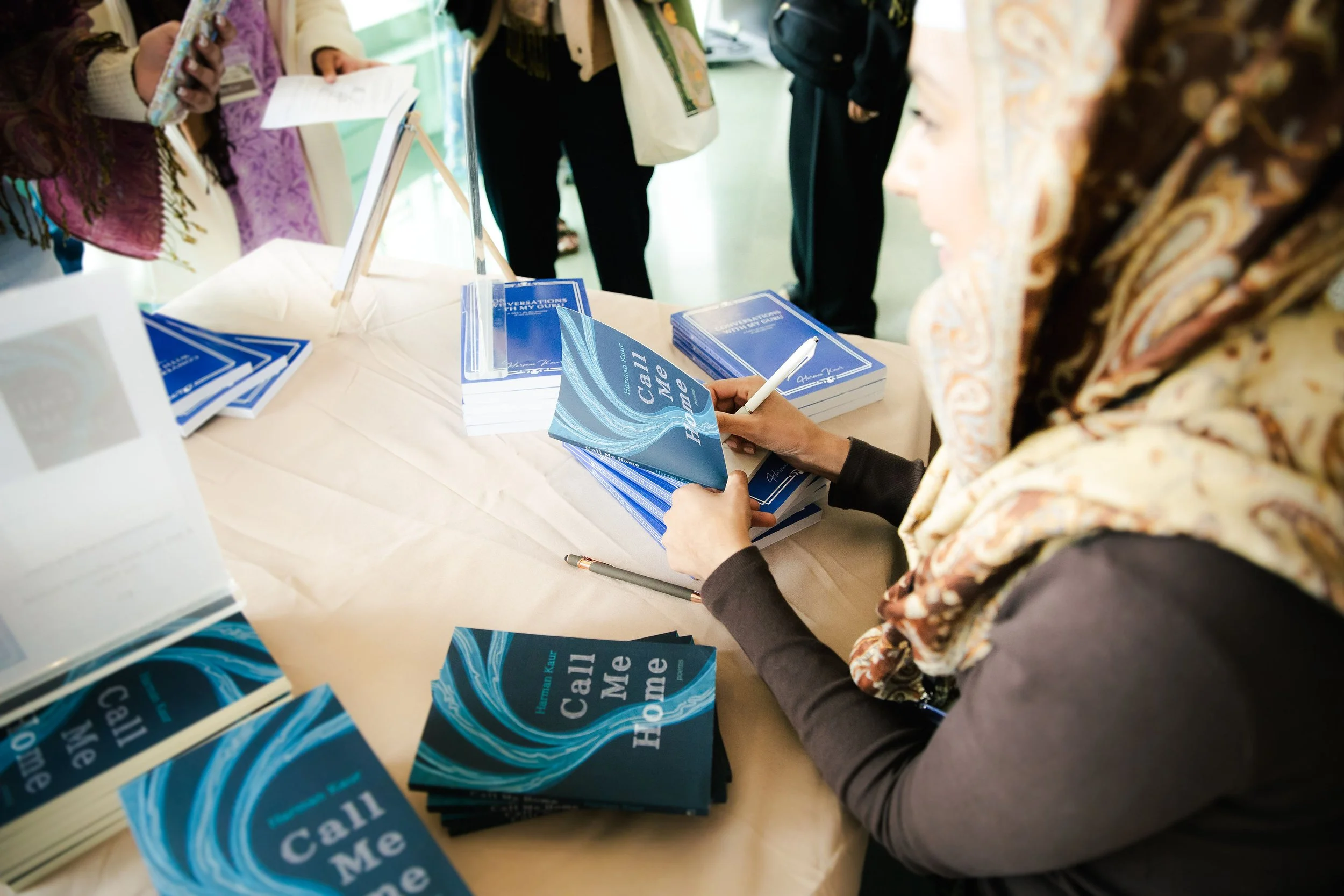 A woman with a headscarf signing a blue book at a table filled with stacks of the same book titled 'Call Me Home' during a book signing event.