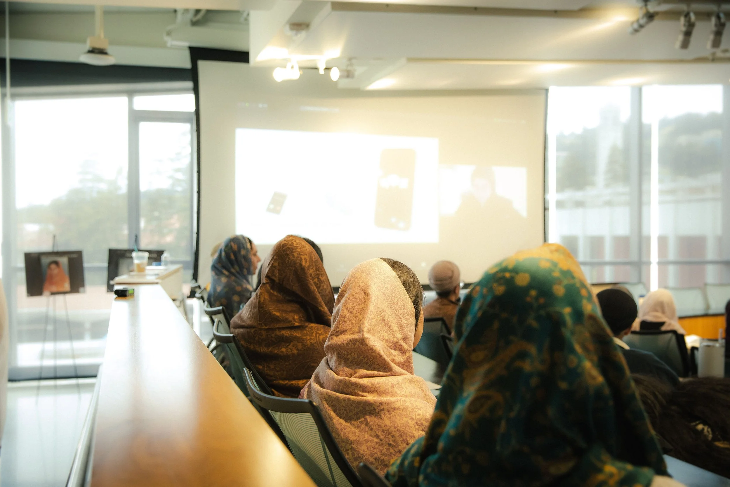 A group of people sitting in a conference room watching a presentation projected on a screen, with some wearing headscarves.