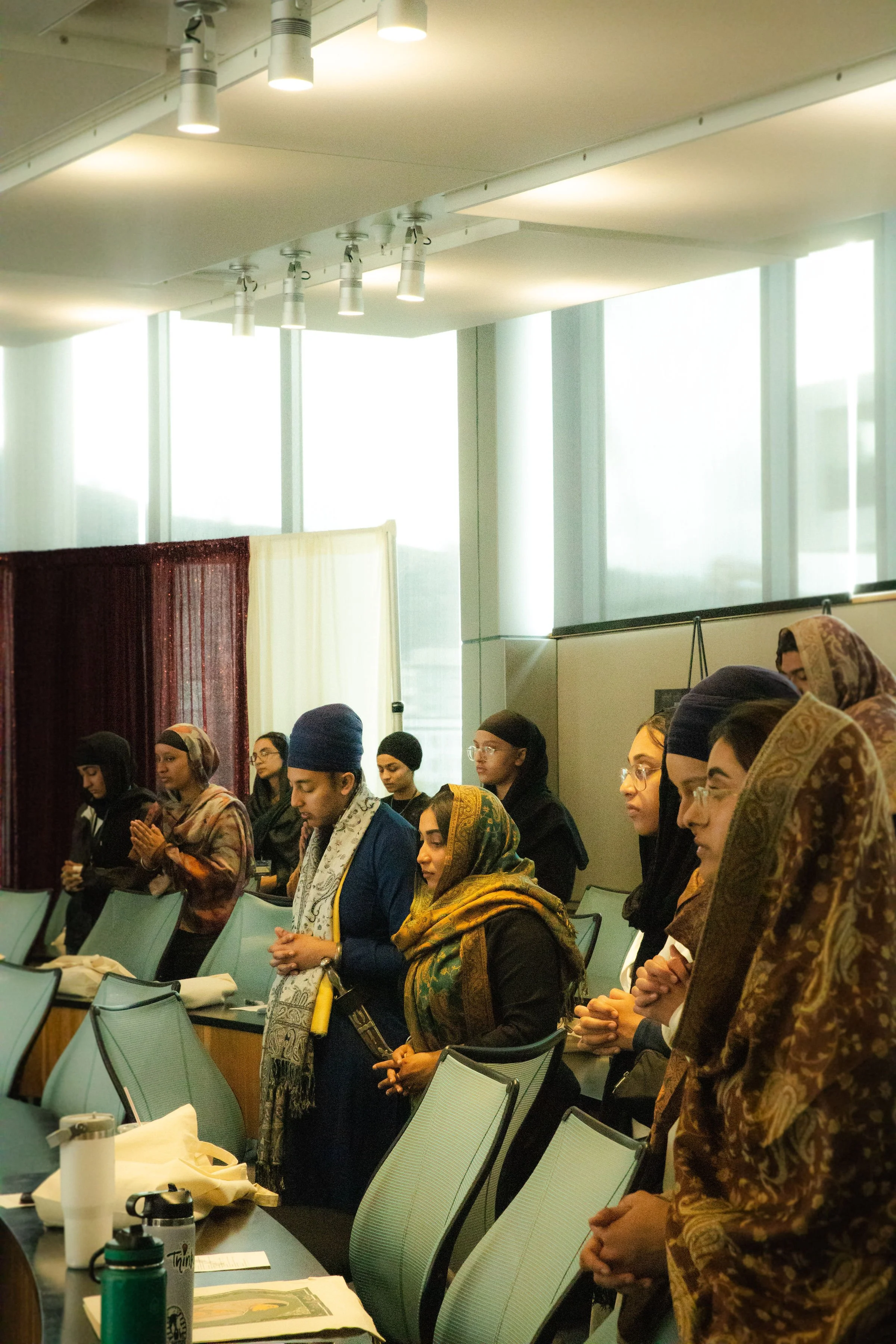 A diverse group of people, mostly women, standing in a conference room with large windows, some with their eyes closed and hands clasped in prayer.