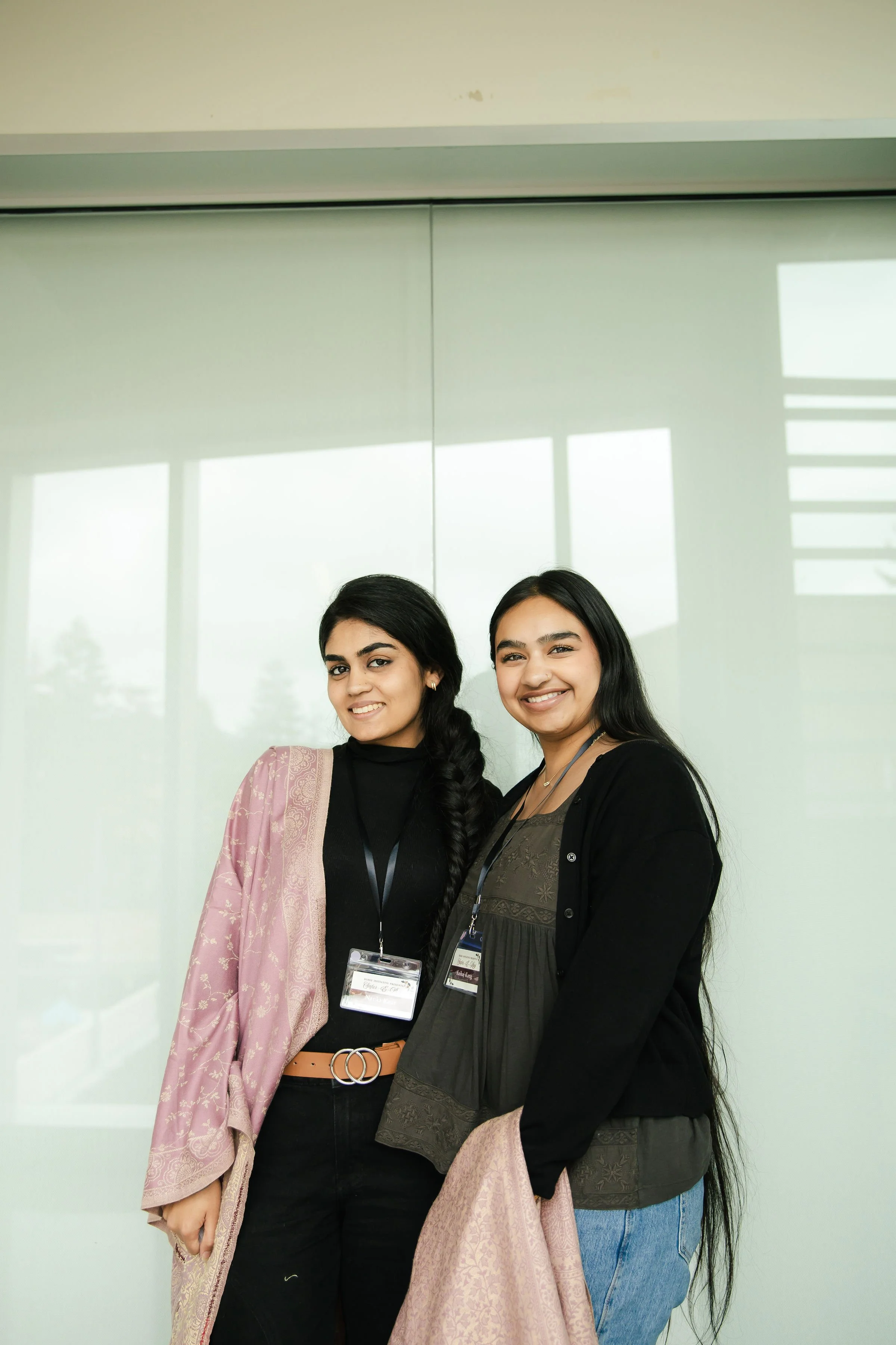Two young women standing side by side smiling in front of a glass wall.