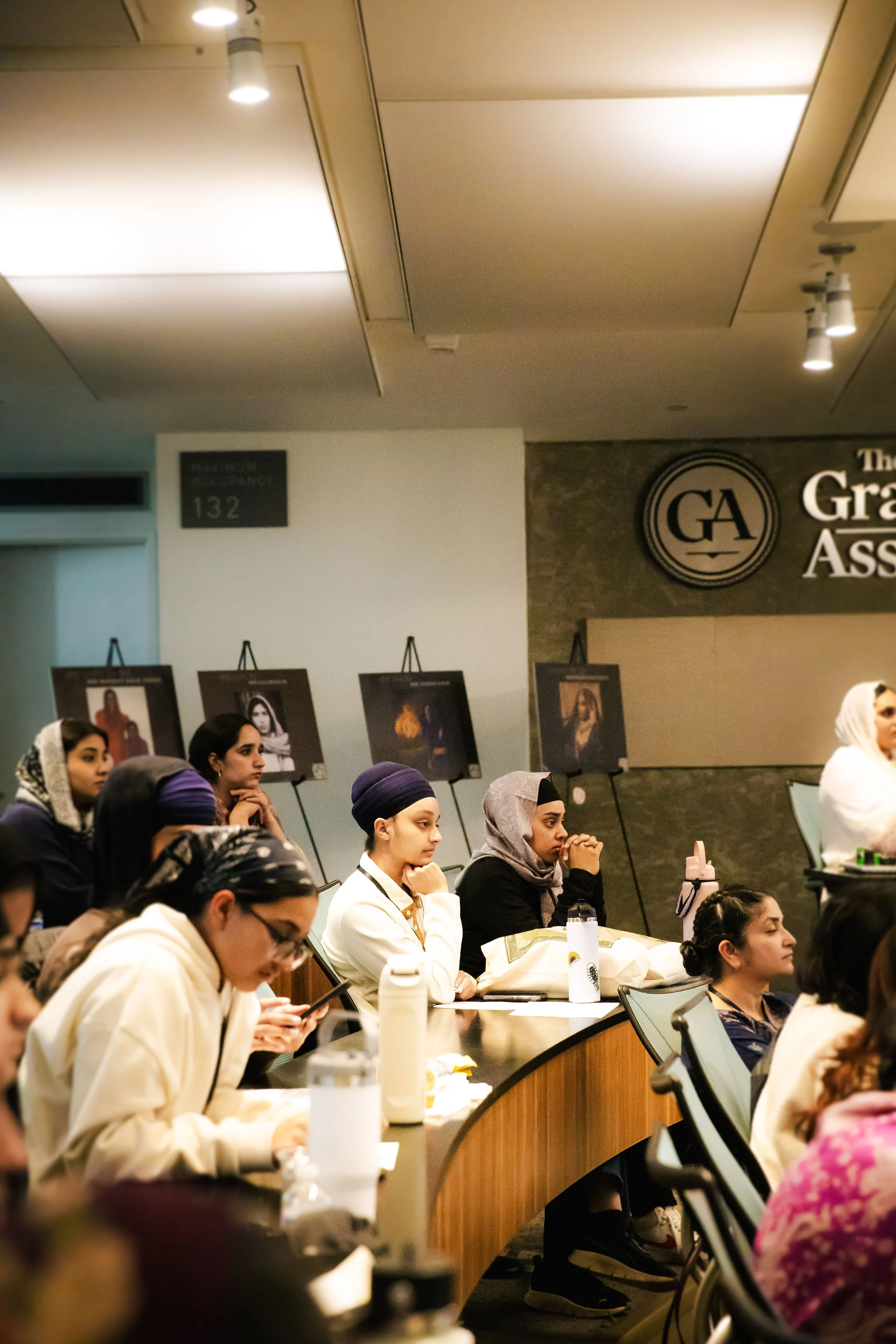 Women attending a conference or seminar, sitting at a curved table, some using phones, with photos on display boards in the background and a sign indicating 'Maximum Occupancy 132'.