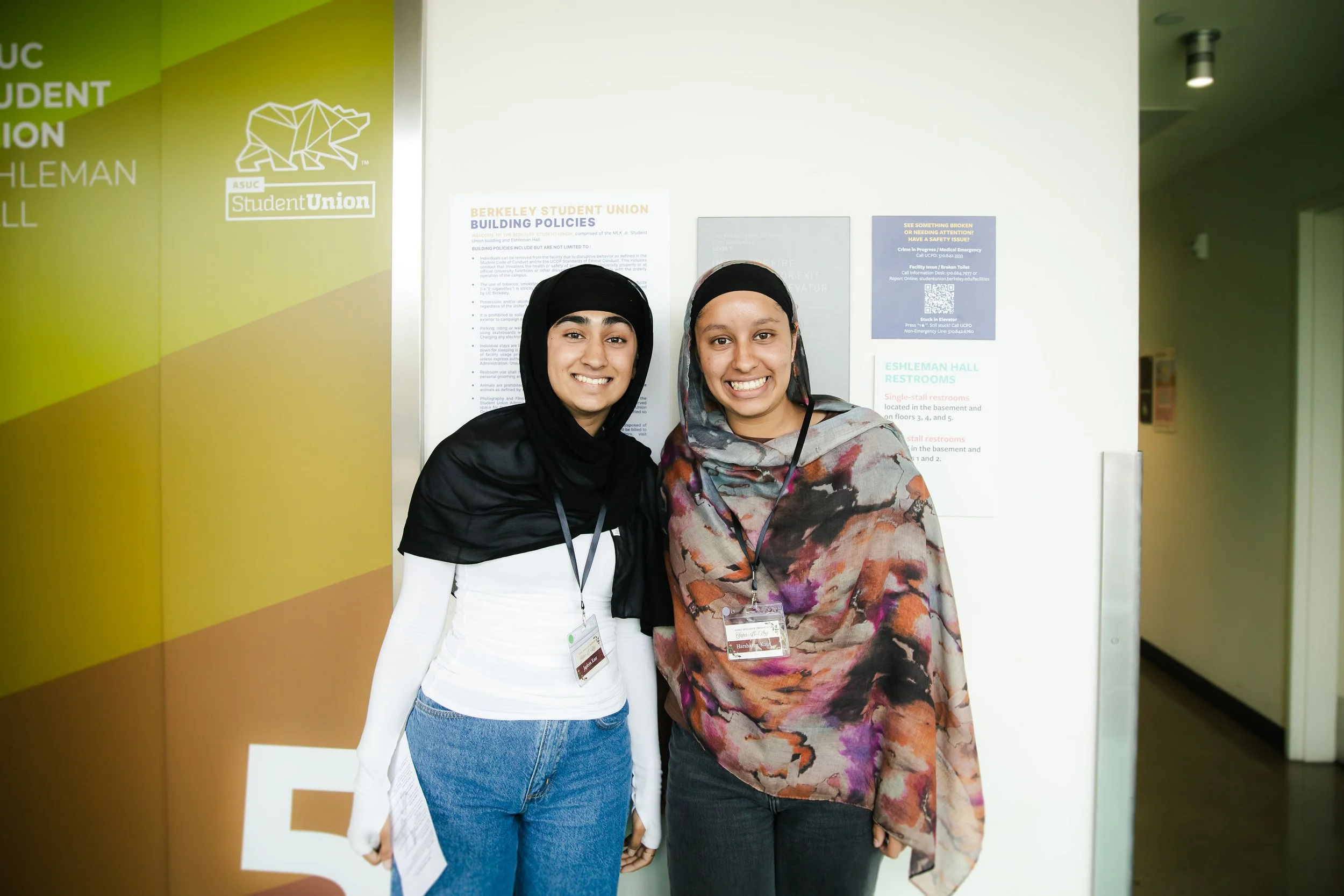 Two women standing close together, smiling, inside a building near a wall with signs and posters.