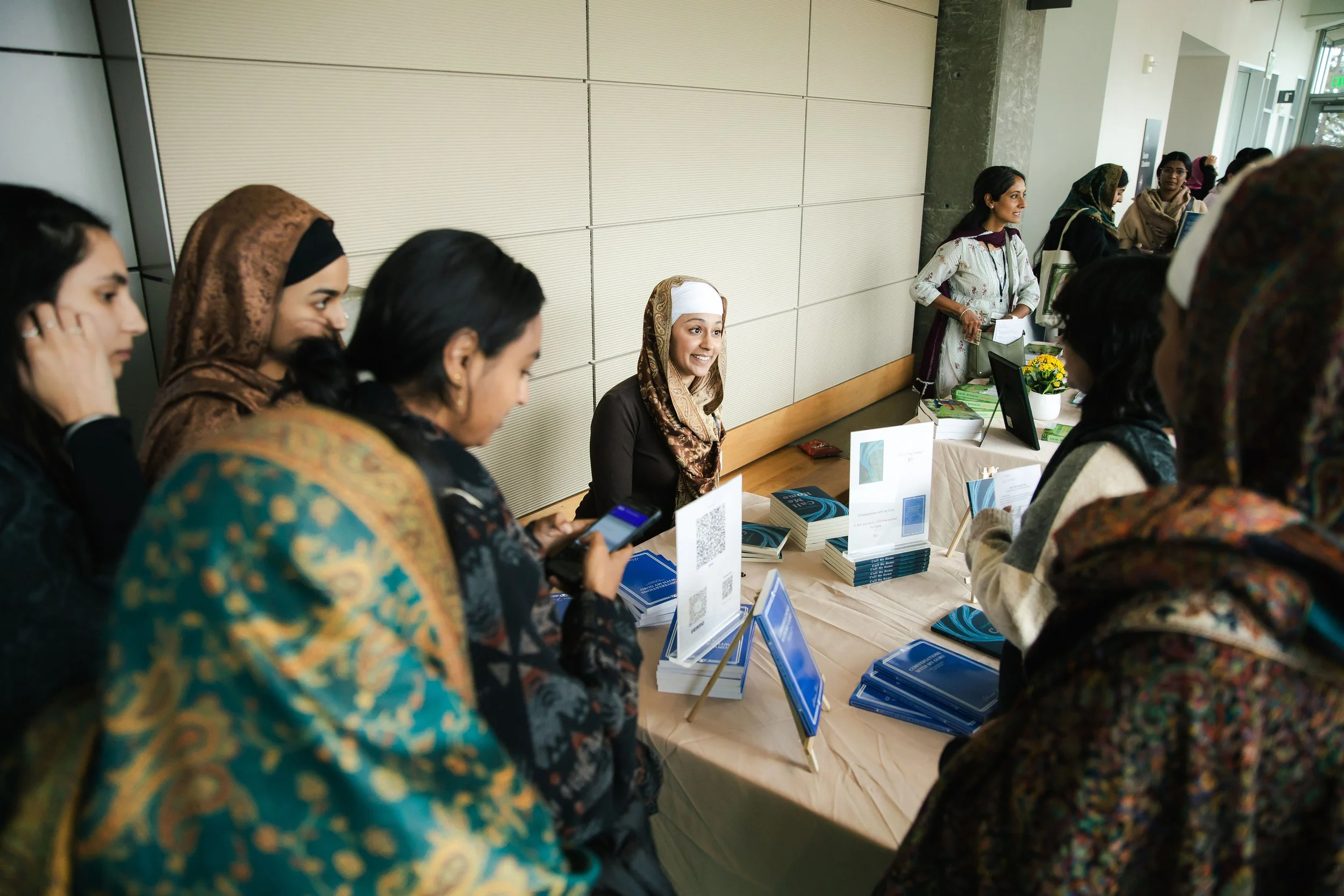 A diverse group of women at a conference or event, with some wearing hijabs. They are gathered around a table with informational materials, brochures, and digital screens, engaging in conversation inside a modern building.
