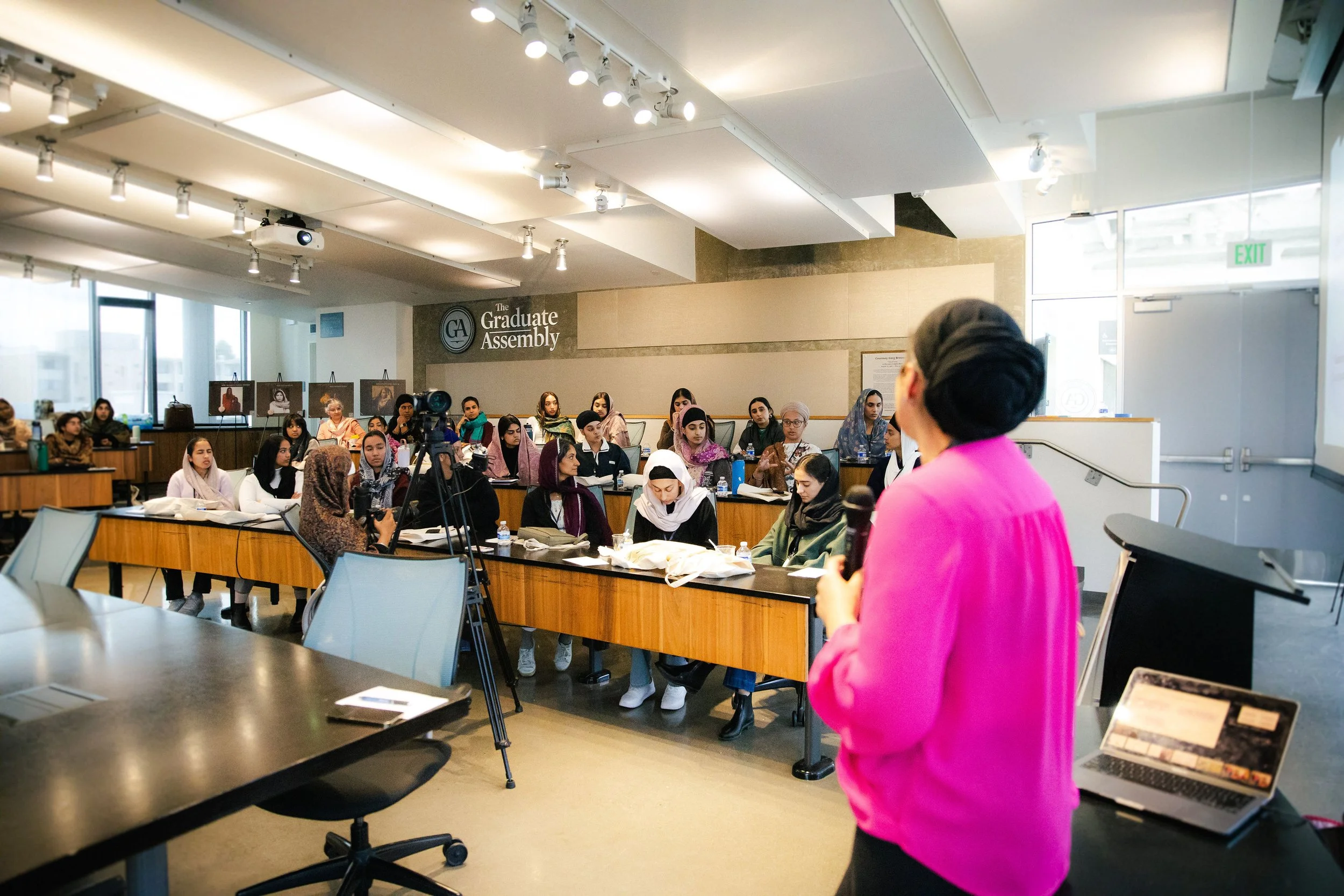 Bibi Supreet Kaur in a pink blazer giving a presentation to a diverse audience in a conference room at The Graduate Assembly, with a camera set up to record the event.